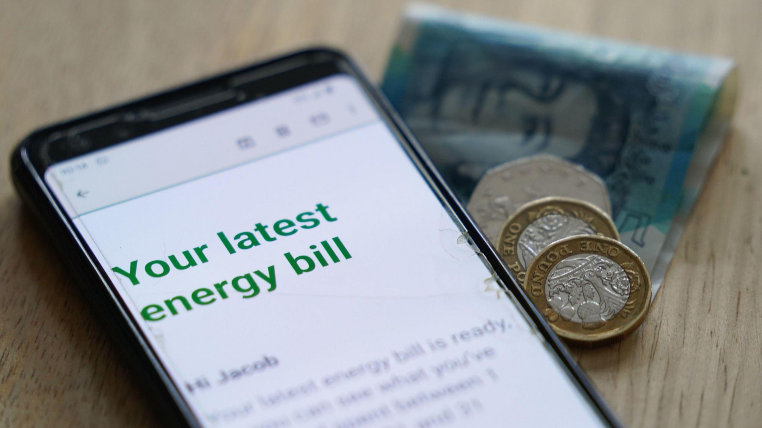 A mobile phone showing a message headed 'Your latest energy bill' and £7.50 in cash are lying on a table.