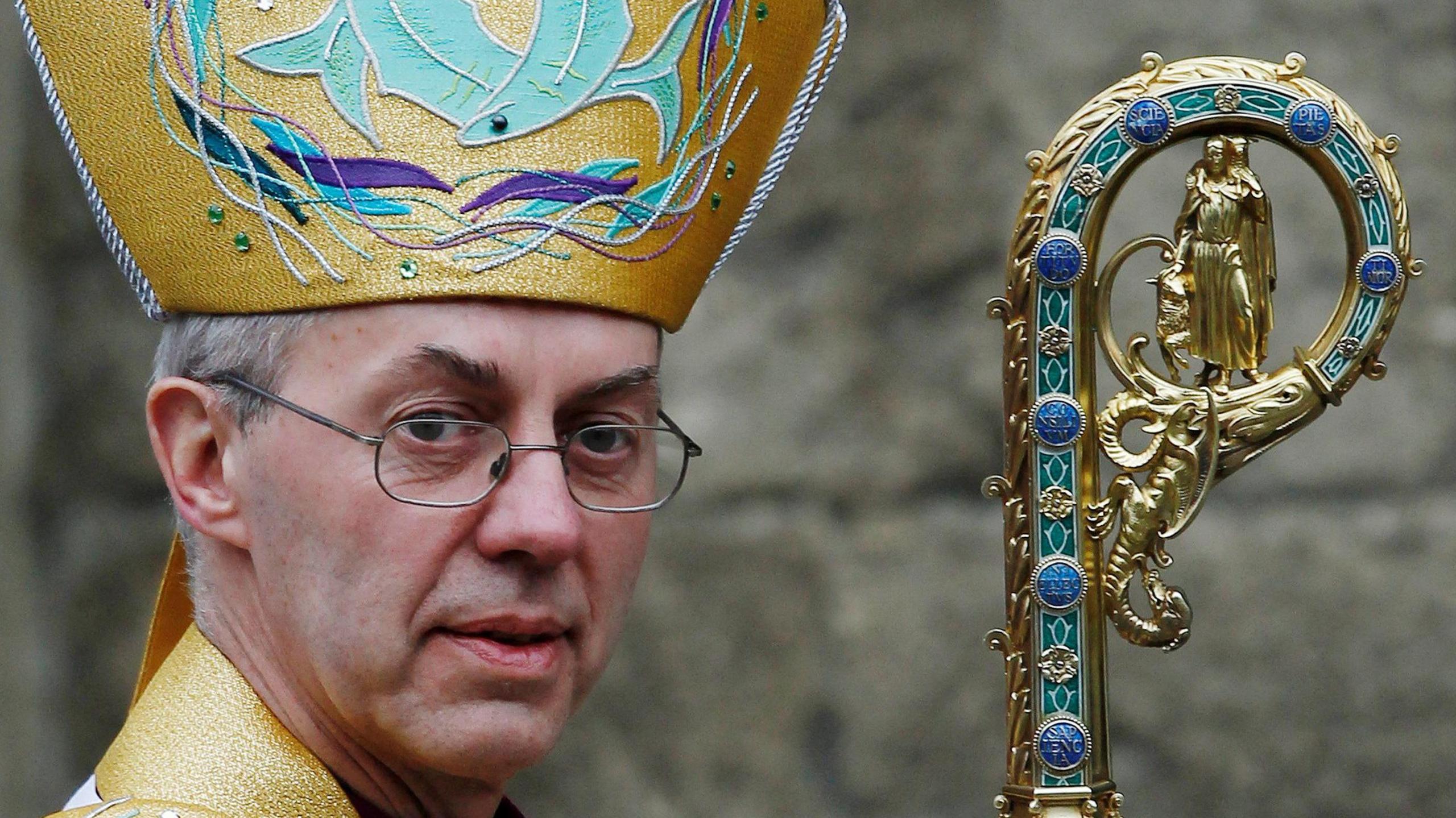 Justin Welby wearing the traditional robes of the Archbishop of Canterbury. 
He is wearing glasses and looking at the camera. He is not smiling.