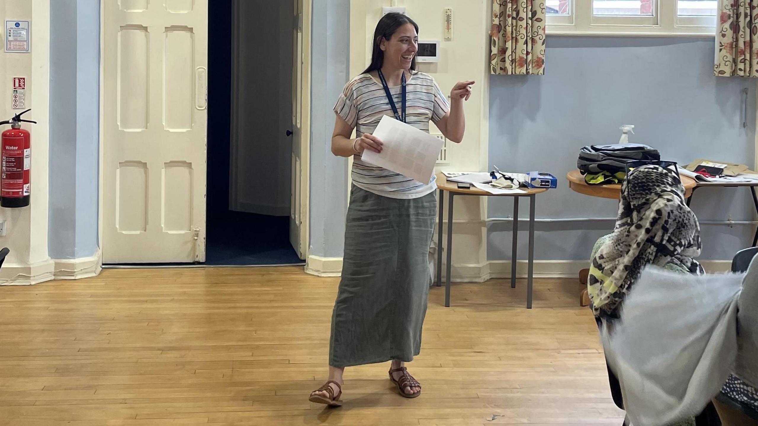 Christie, a woman, hosting a language class, smiling and pointing her finger. She has long dark hair and is wearing a stripy top and  grey ankle-length skirt with brown, Grecian sandles 