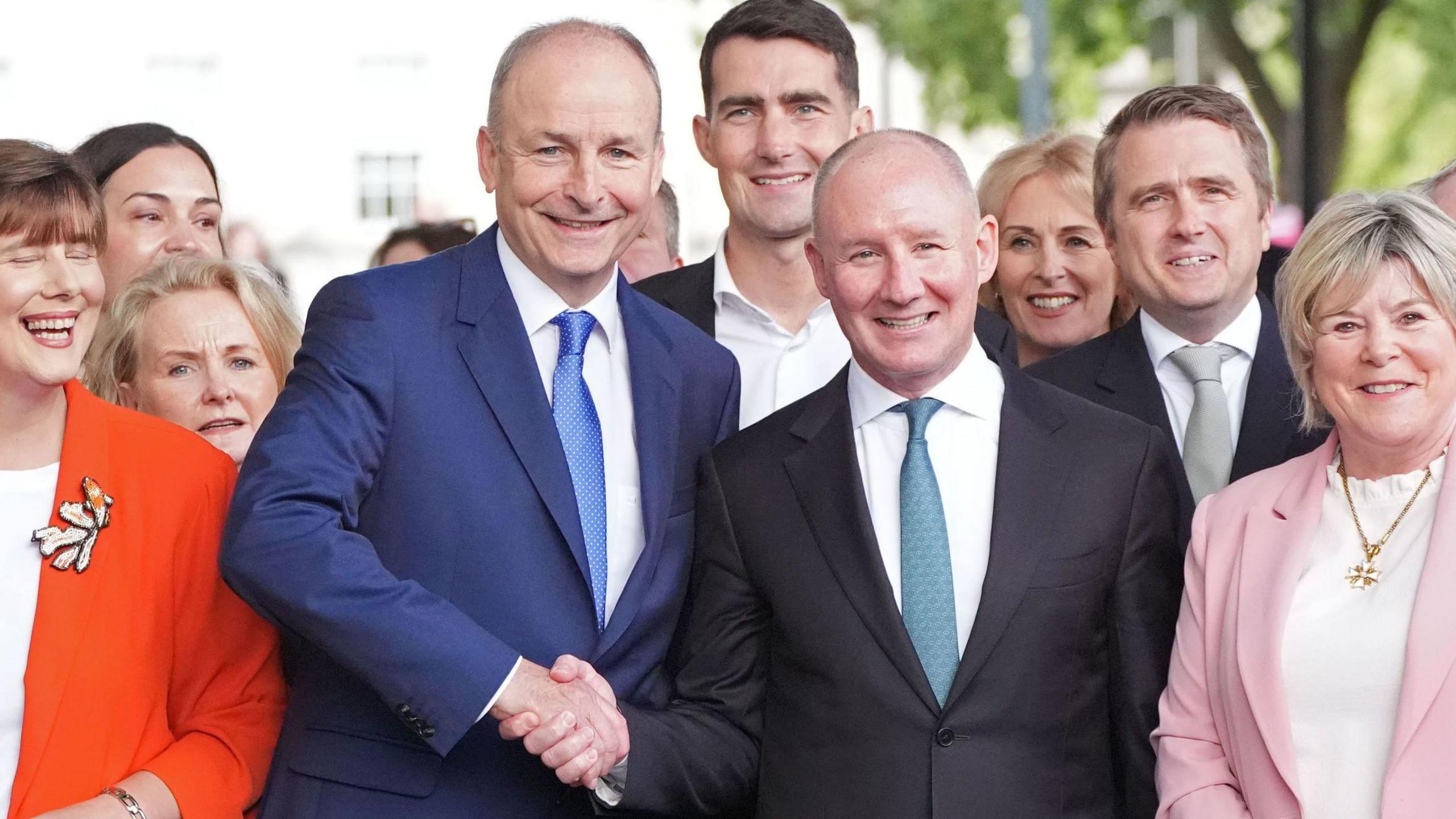 Micheál Martin shakes hands with Jim Gavin and smile at the camera as they stand among other Fianna Fáil party colleagues. Martin and Gavin are both older men with balding/greying hair. Martin is wearing a navy suit jacket, a blue tie and a white shirt. Gavin is wearing a black suit jacket, a light teal tie and a white shirt.
