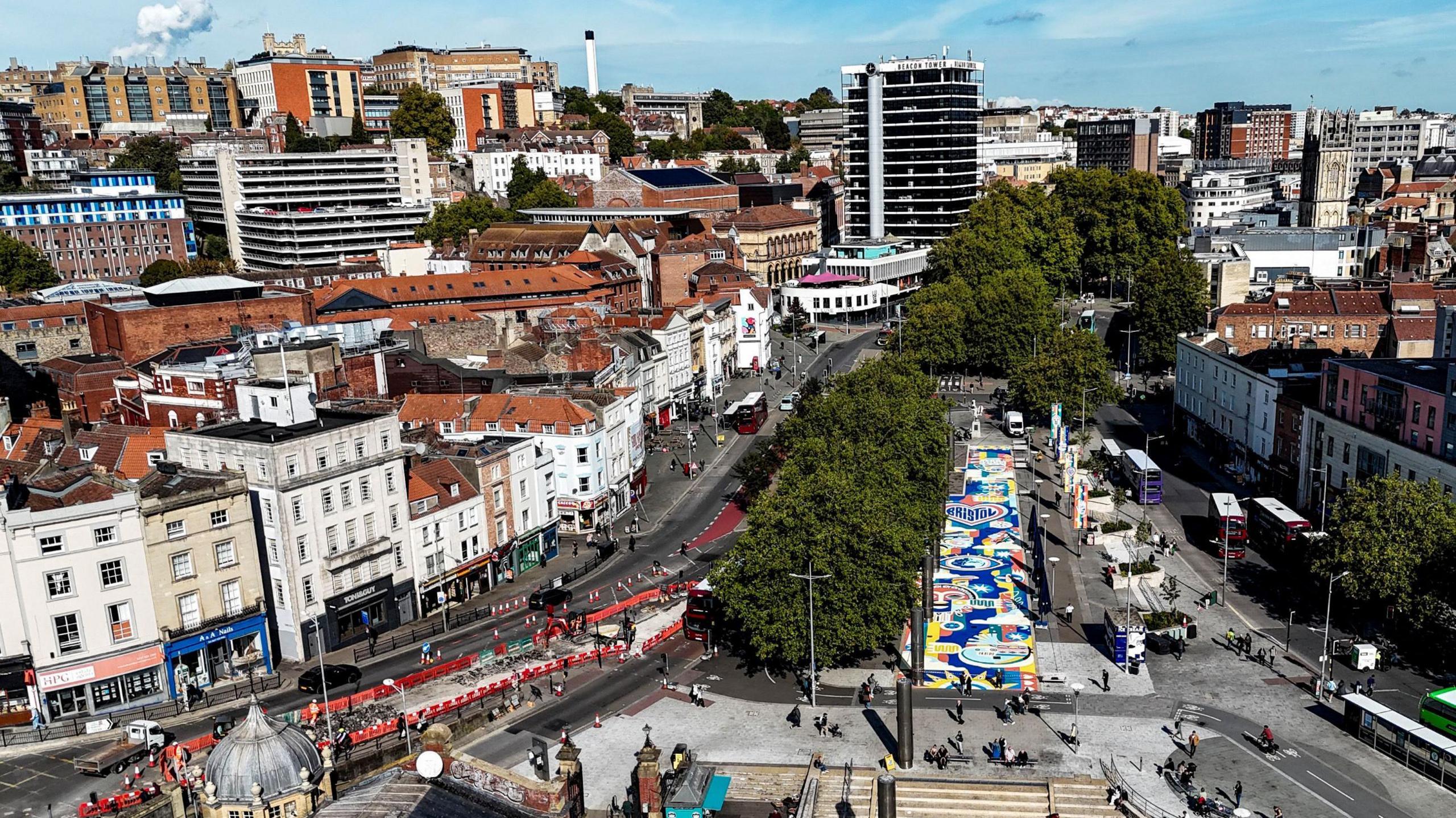 A view taken from a drone of Bristol city centre showing the area between the Hippodrome and the harbourside from high up. At the bottom right-hand side of the picture is a new mural called Our Common Ground which is a combination of different shapes and letters, mostly coloured in yellow and blue.