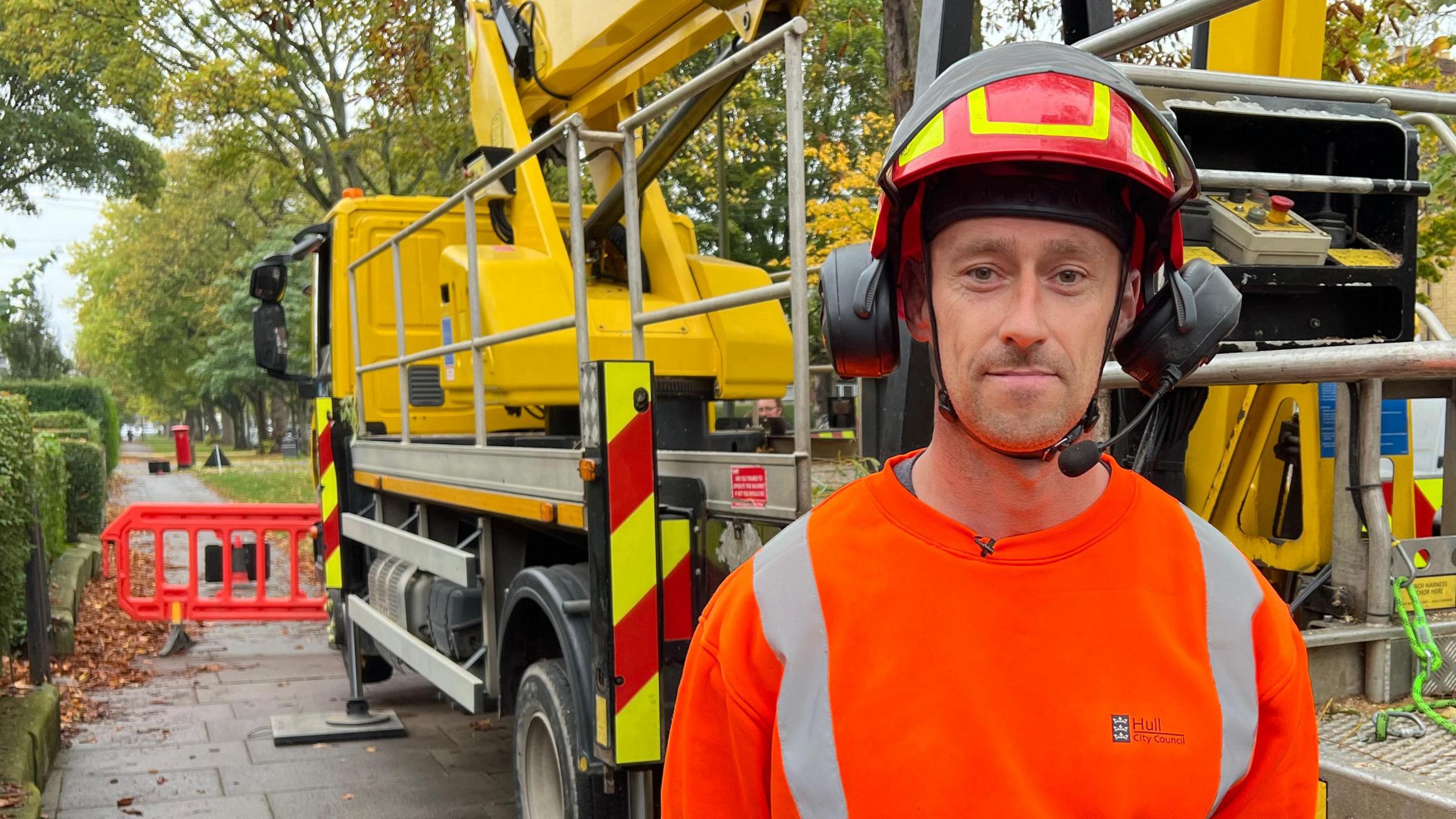 Tree surgeon Craig Davis wears a red and yellow helmet, ear protectors and an orange, hi-vis jumper. He is standing in front of a crane on the back of a truck, which is parked on the pavement in a leafy Hull avenue. A red barrier blocks the path and green trees can be seen.