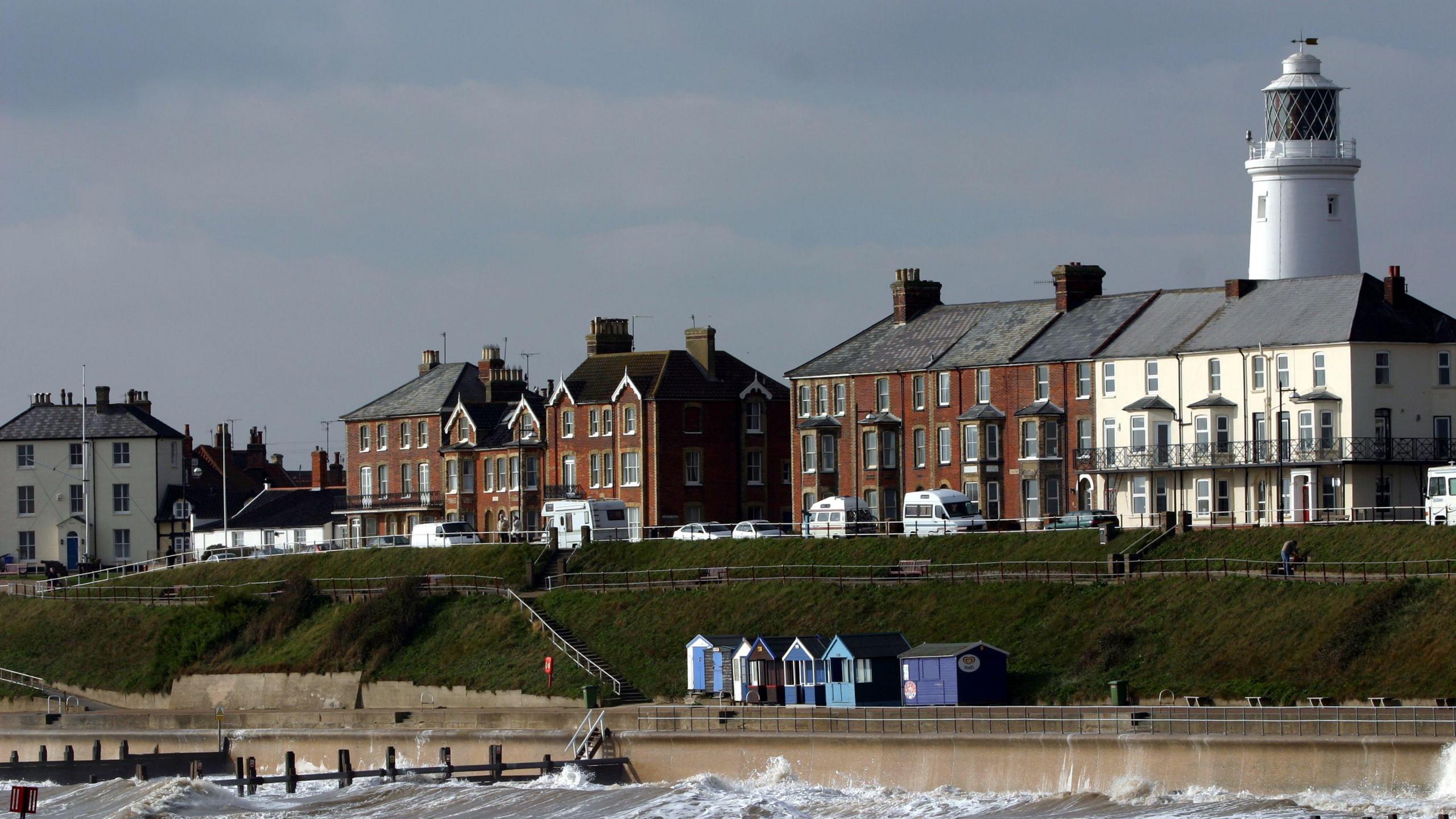 The Southwold coastline. Homes overlook the sea with cars parked outside of them. A white lighthouse can be seen behind them. Beach huts and a high sea wall sit in front of the sea.