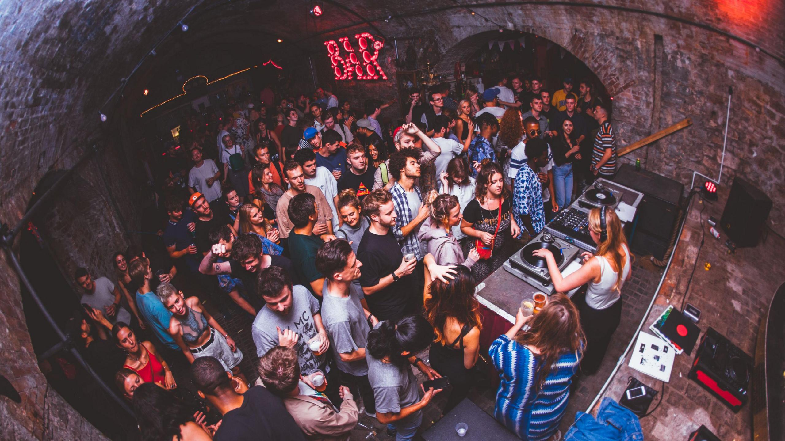 A large group of people dance inside Bristol underground venue Loco Klub. The internal walls are made of large dark brickwork and a female DJ is playing on a set of decks at the front of the crowd