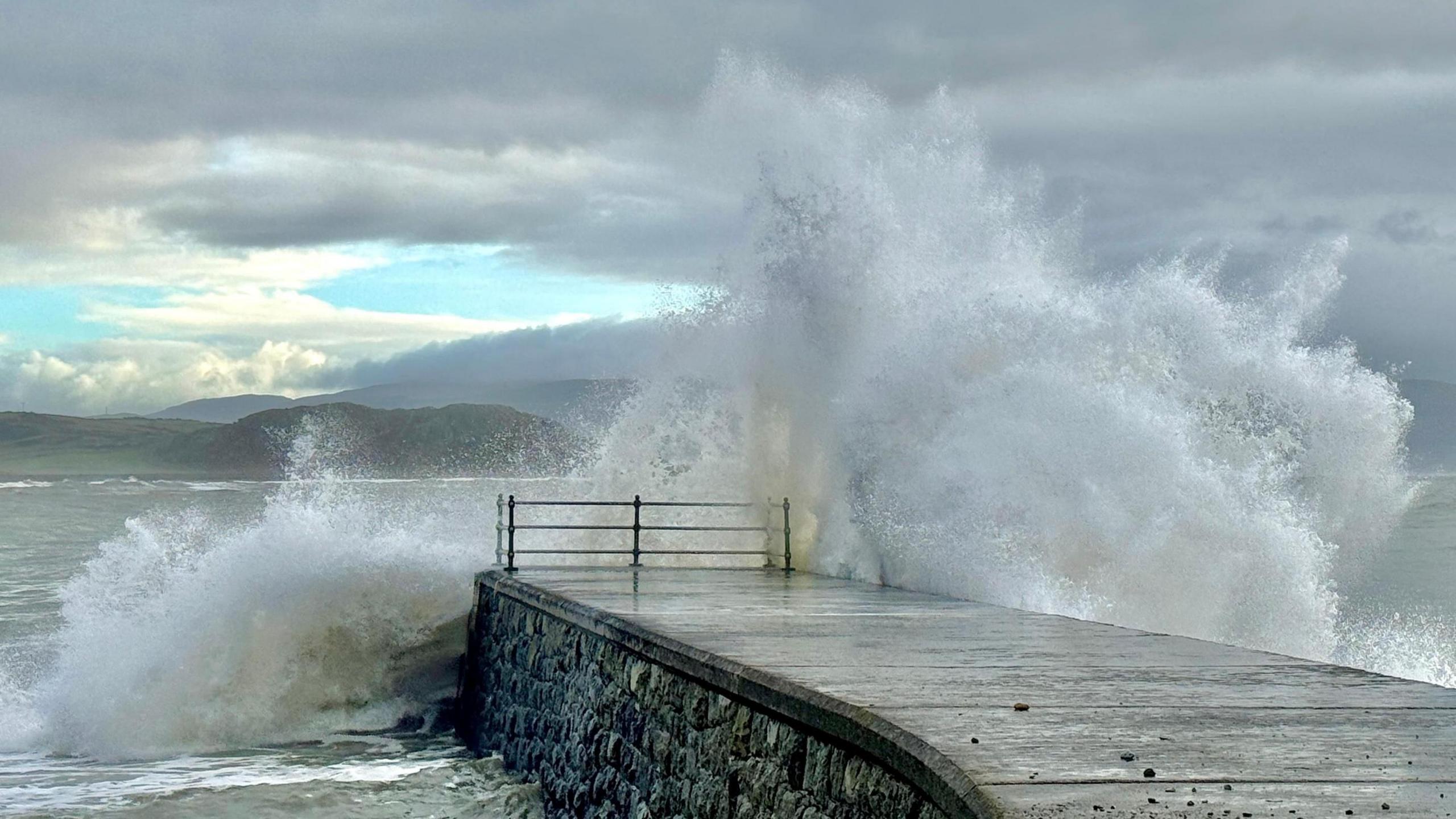 A wave crashes over a sea wall with hills in the background and a mostly cloudy sky overhead