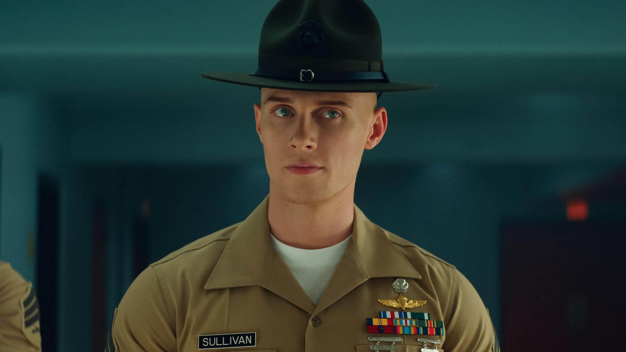 Max Parker as Sgt Sullivan smirks in a still fromthe show. He is in military gear with a brimmed dark hat and a khaki green shirt with a set of medals and name badge on his lapels.