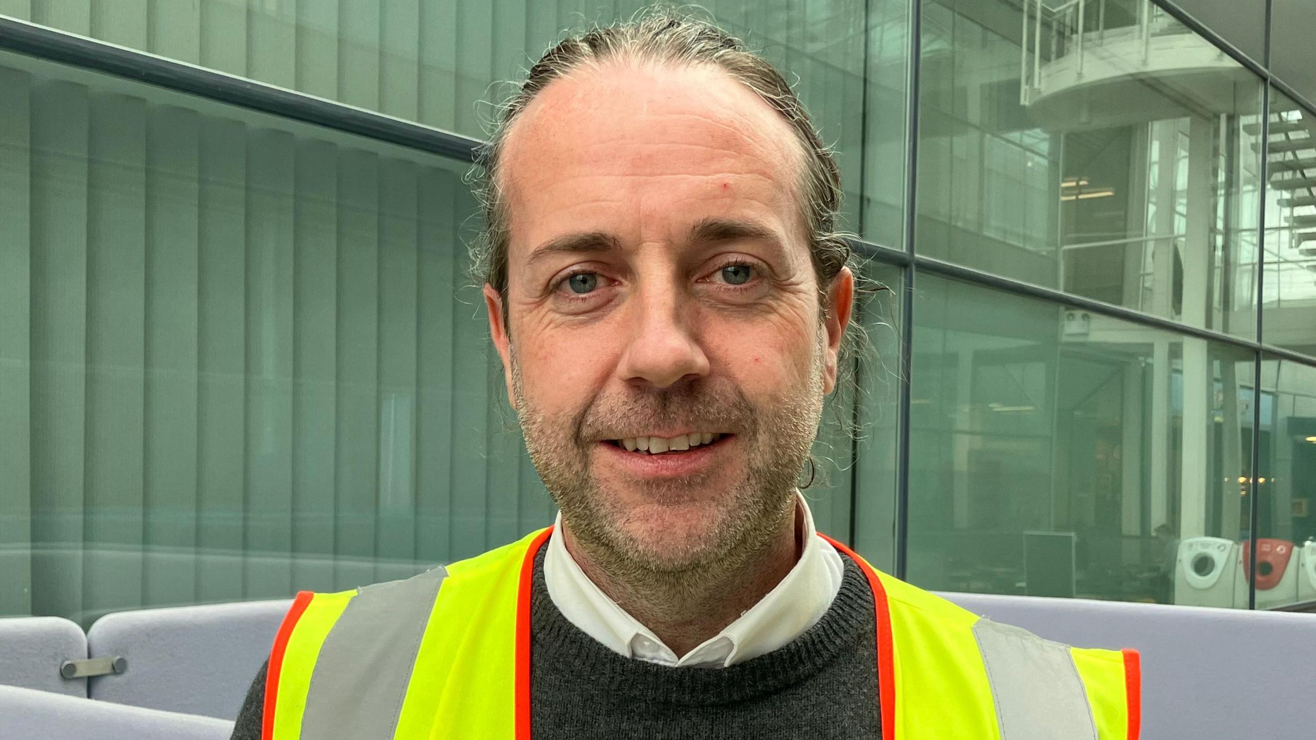 A head and shoulders picture of Colin Walsh wearing a high-vis jacket inside a building at Stansted Airport. His hair is swept back and he has facial stubble. He is wearing a white shirt under a grey crew-neck jumper.