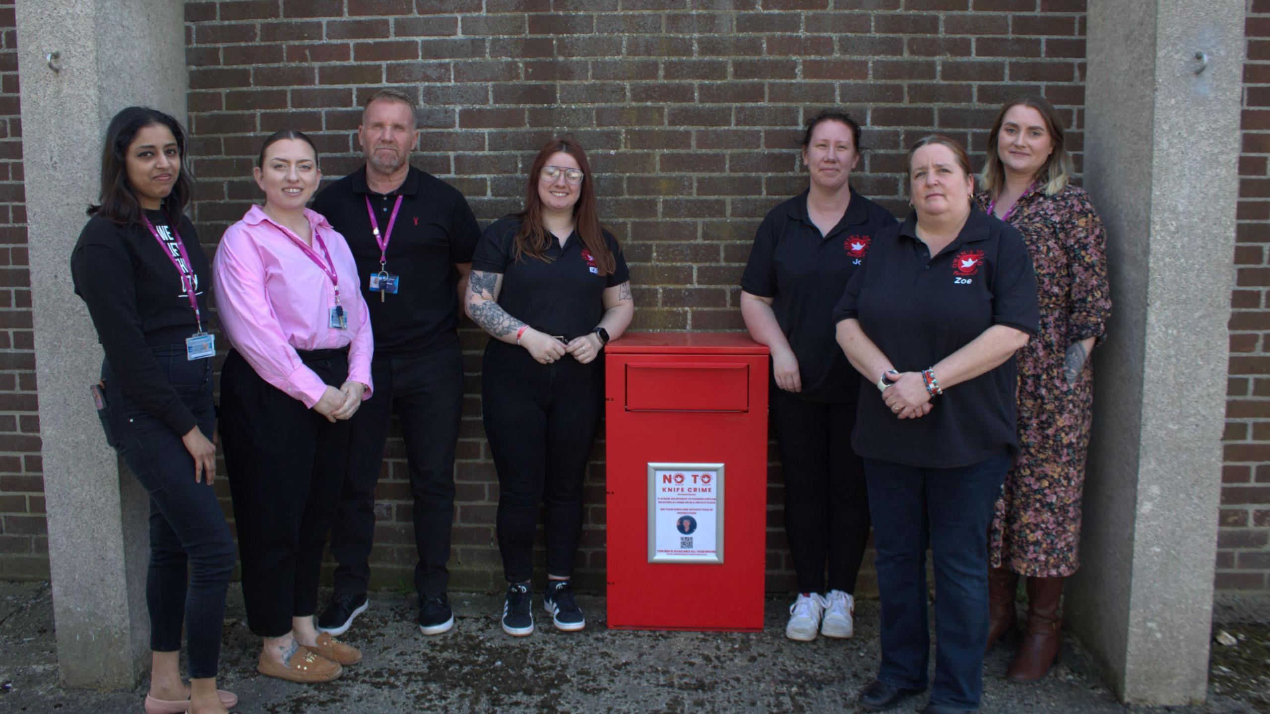 Several people look at the camera next to a brick wall, which in the middle of them has a red metal container attached to it. It has door or drawer at the top that knives can be put in to