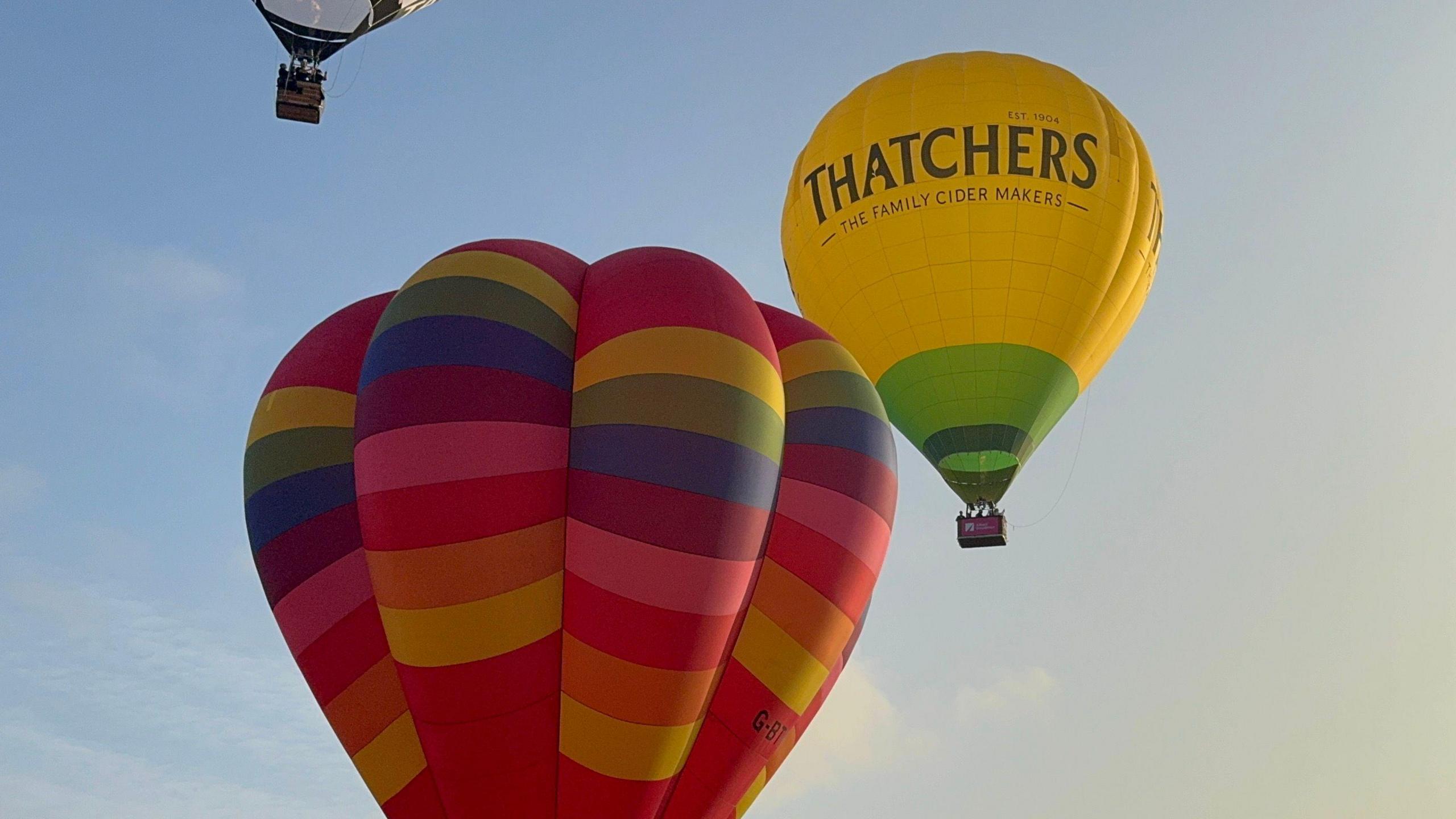 Three hot air balloons in a hazy yellow and blue morning sky. One is rainbow striped and one is a yellow balloon with the Thatchers cider logo on it in green letters. A third balloon is just out of frame, but the basket full of passengers can be seen.  
