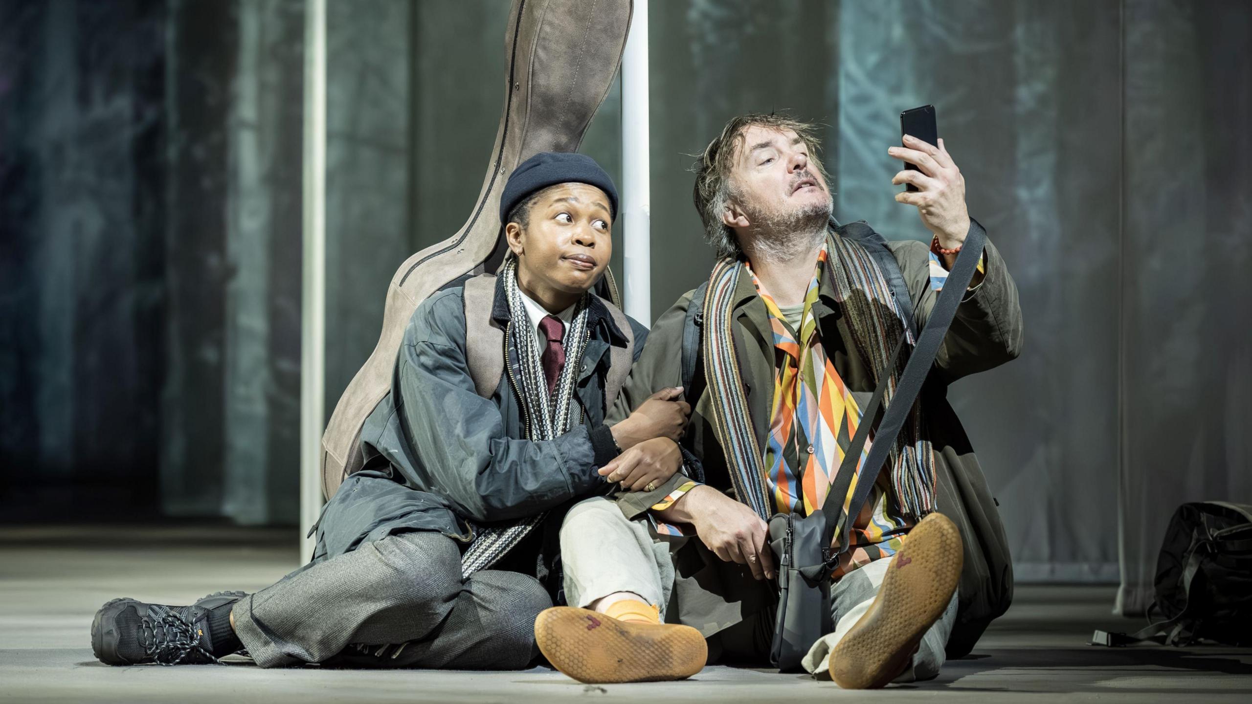 Two actors, one male and one female, lean on each other as they sit on a theatre stage. They are both wearing scruffy clothes. The woman has a guitar in a case behind her, and the man is studying a mobile phone he is holding in the air