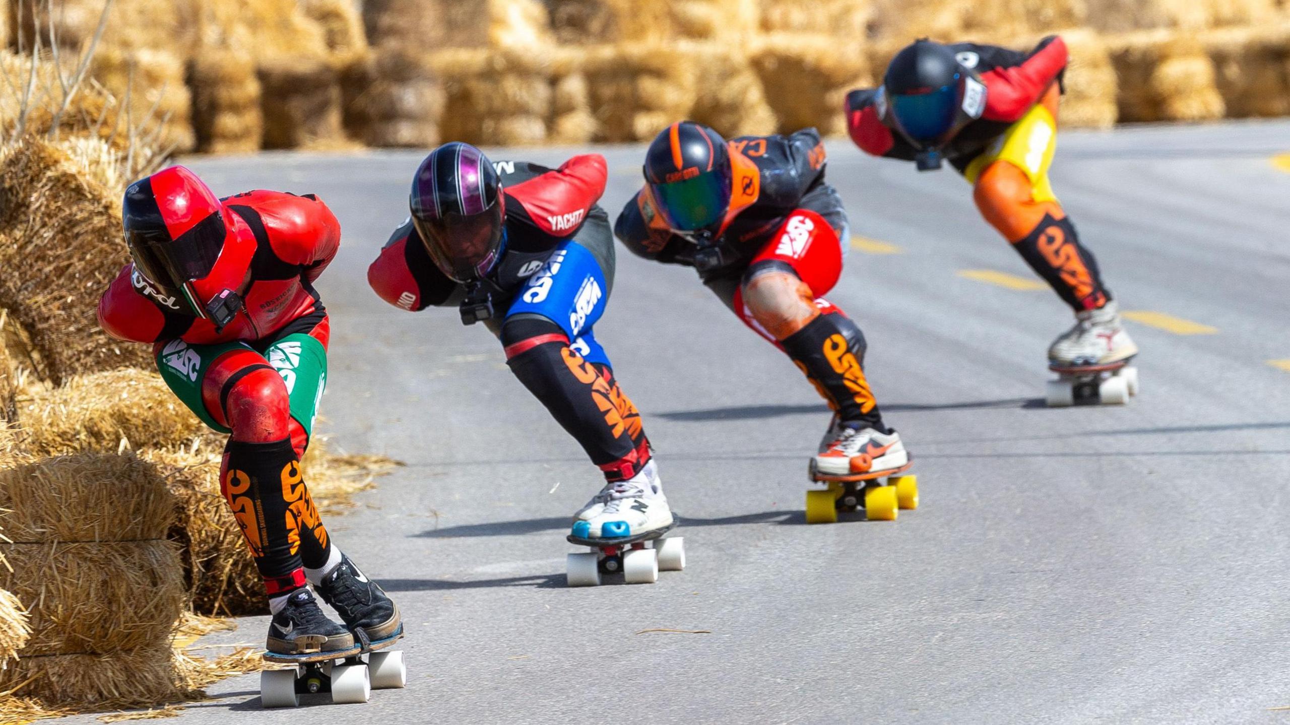 Four people in brightly coloured leathers and helmets on skateboards bending down to lean into a corner on a road. Yellow hay bales line the course.