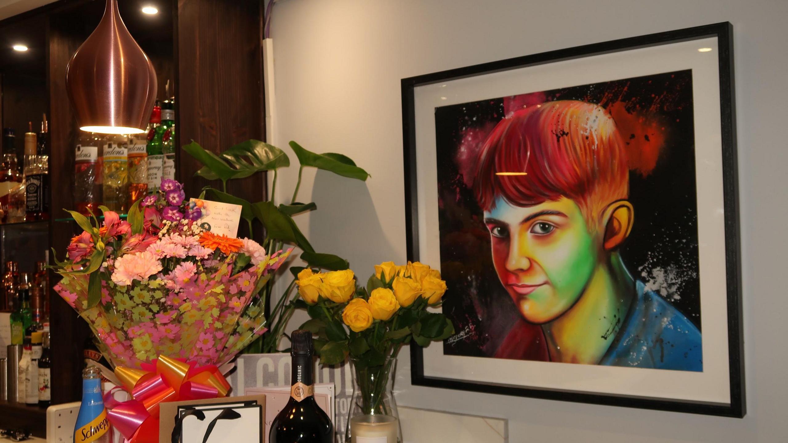 A multi-coloured illustration of a boy in a black frame hangs on a white wall near a bar. Two bouquets are placed on the bar.