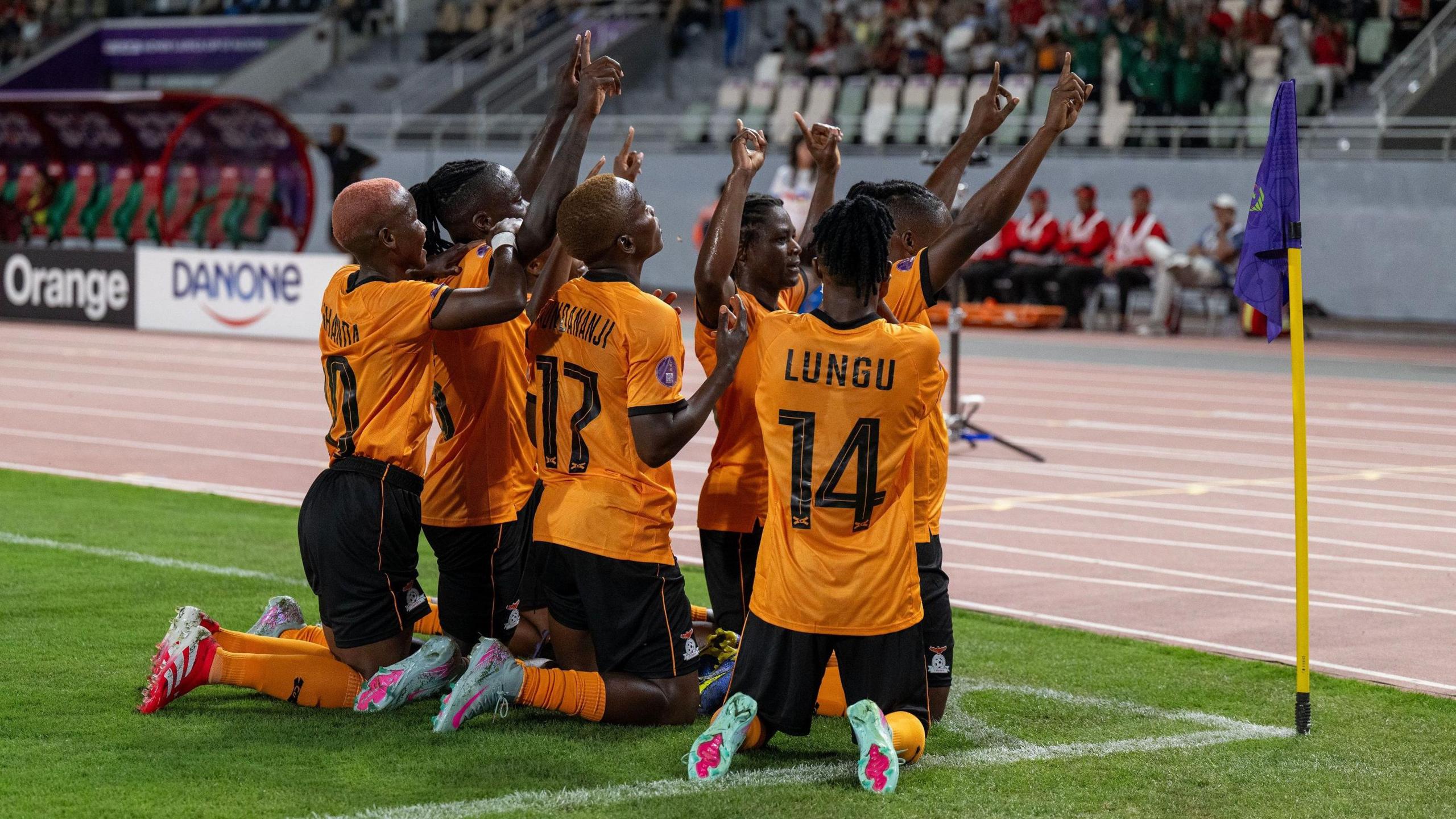 Zambia players kneel down in front of a corner flag and raise their arms in celebration after scoring against Morocco, with a running track seen around the edge of the pitch near them