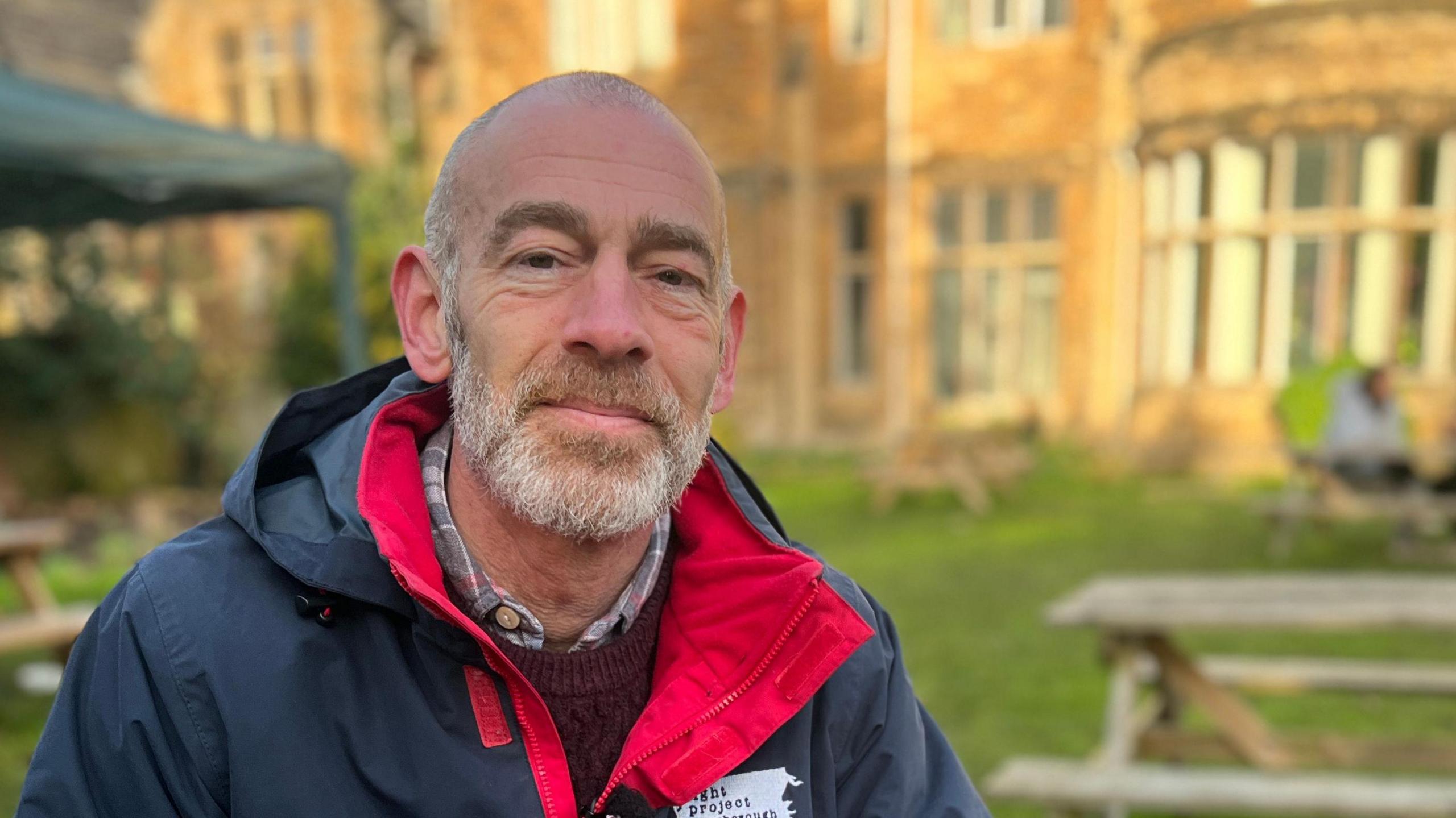 Steven Pettican is sat on a picnic table outside the Light Project building and smiling. He wears a branded navy coat with red on the inside, a red jumper and checkered shirt. He has grey short hair and a grey beard. 