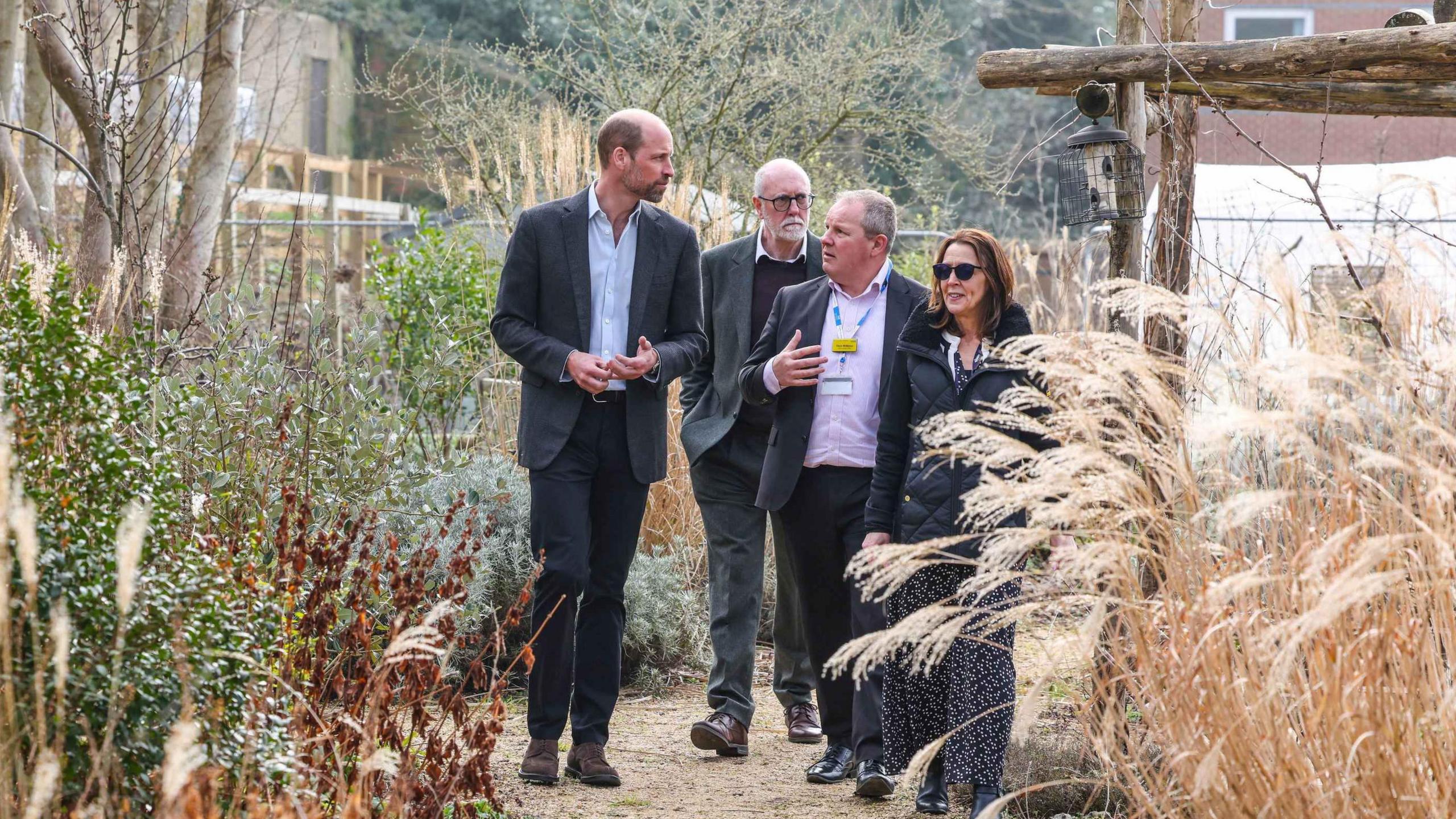 The Prince of Wales, patron of NHS Charities Together, walking in a garden with two men and a woman. There are tall ornamental grasses at each side of the path they are walking along. They are walking towards the camera.