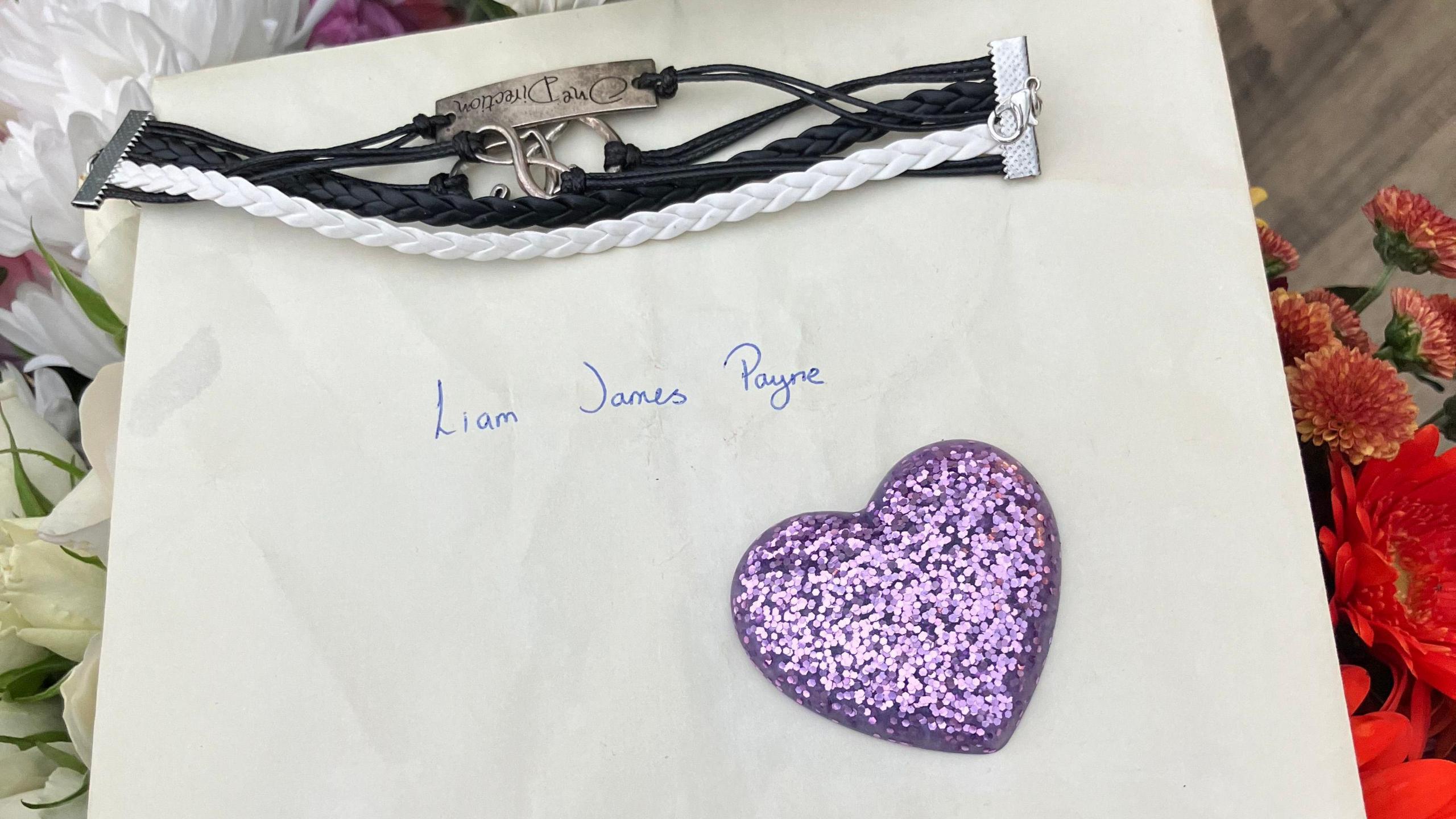 A card addressed to Liam James Payne of One Direction. A purple glittery heart is on the envelope, as well as a white and black piece of jewellery. Flowers can also be seen in the photo. 