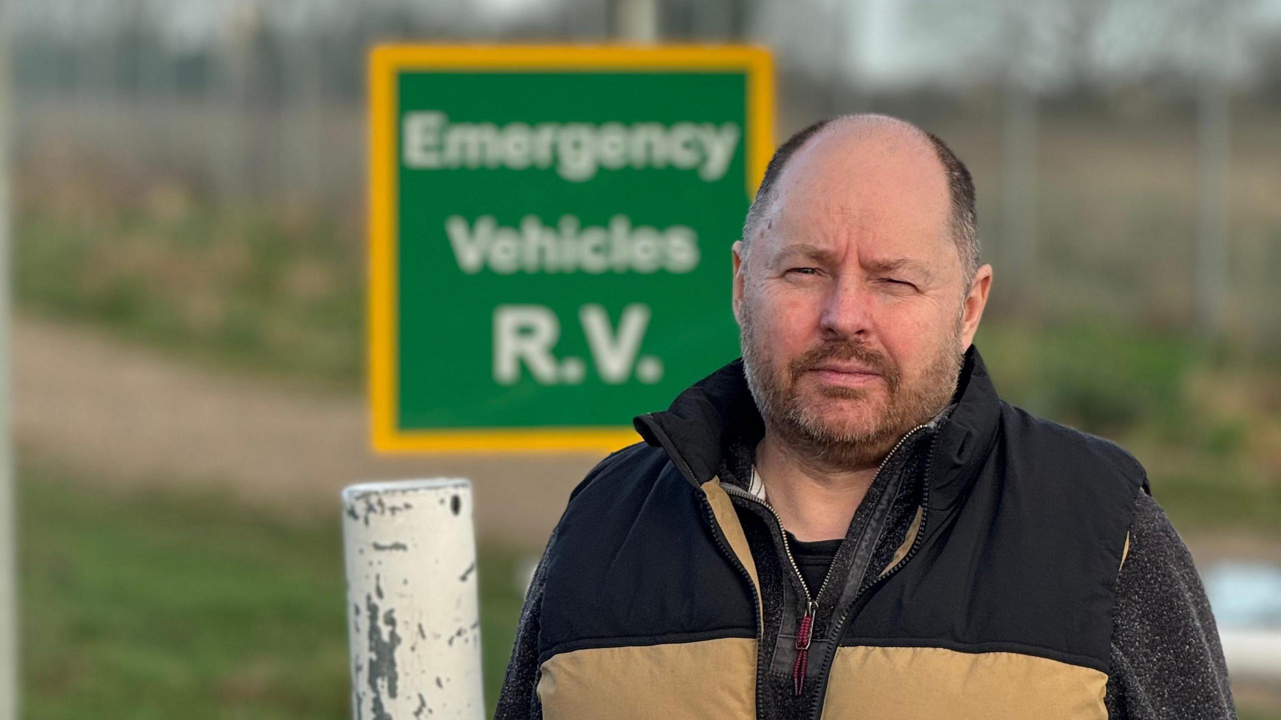 A bald man wearing a black and sand-coloured Gillet over a grey jumper standing in front of a green sign which reads Emergency Vehicles R.V