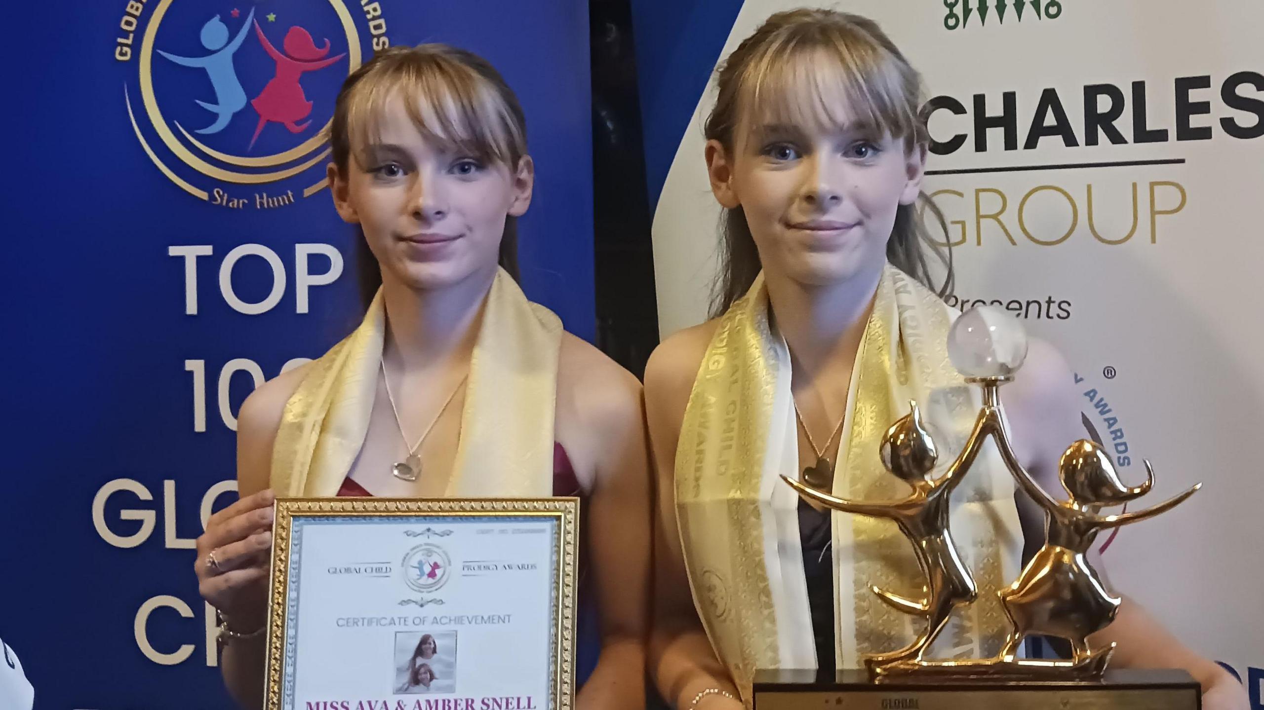 A pair of teenage girls who are identical set of twins, both blonde and wearing gold scarves over their necks, holding up a gold award and a framed certificate.