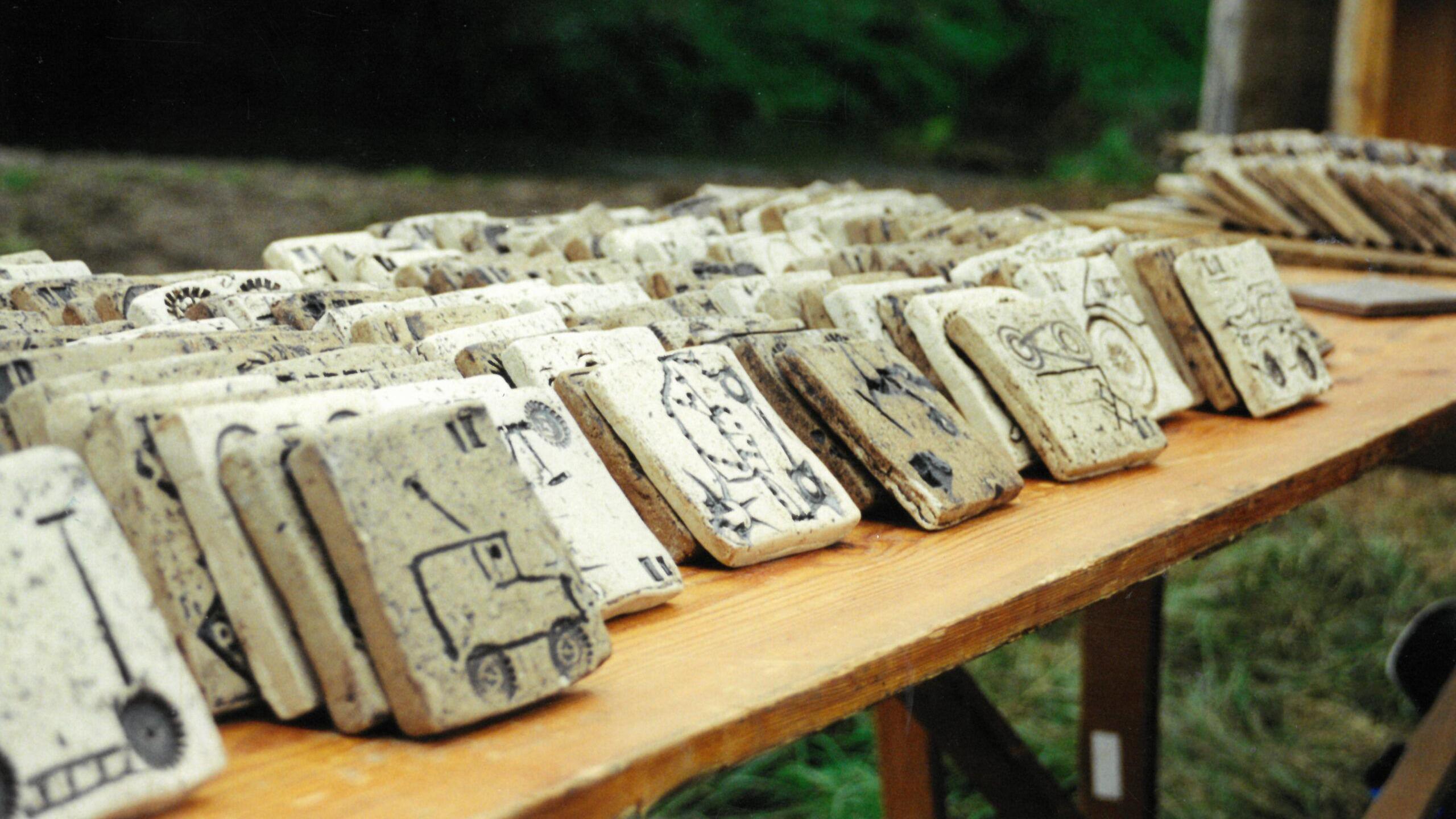 Stacks of tiles leaning against each other on a table outside, each with a carefully etched unique design, like black pencil drawings.