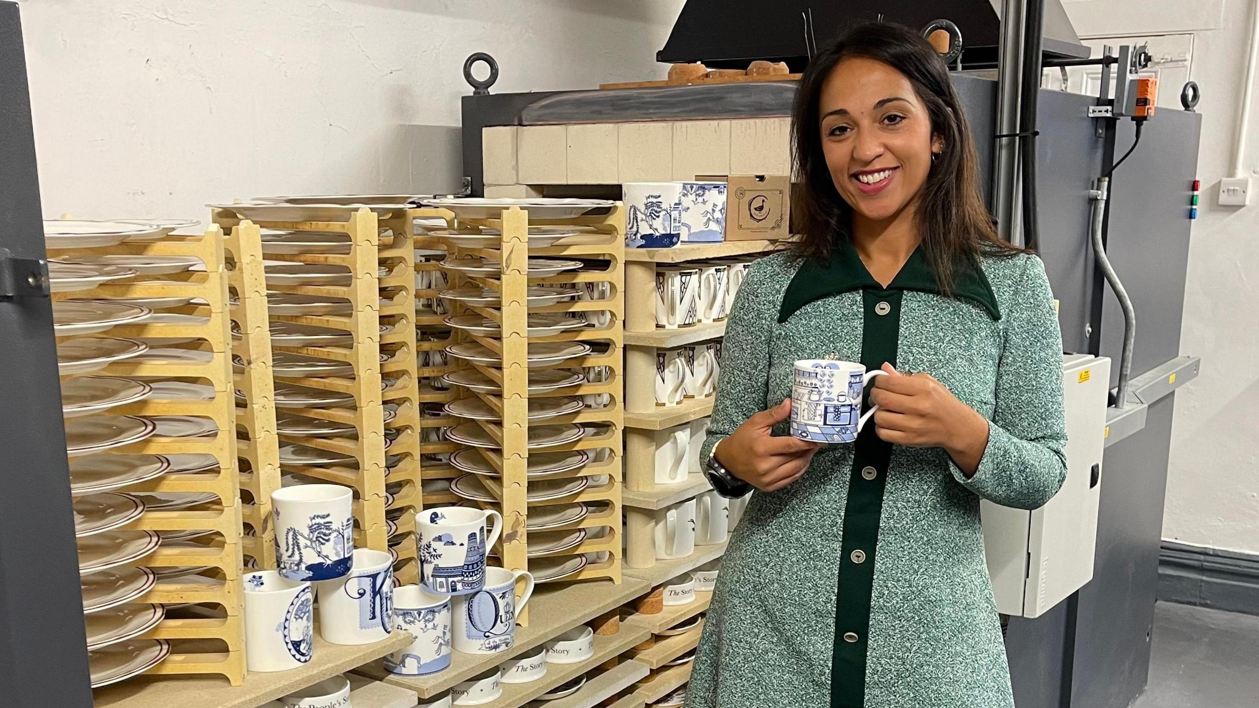 A woman with shoulder length dark hair stands beside a modern pottery kiln. The kiln is open and filled with decorative china plates and cups. The woman is holding one of the cups which has a blue and white design. 