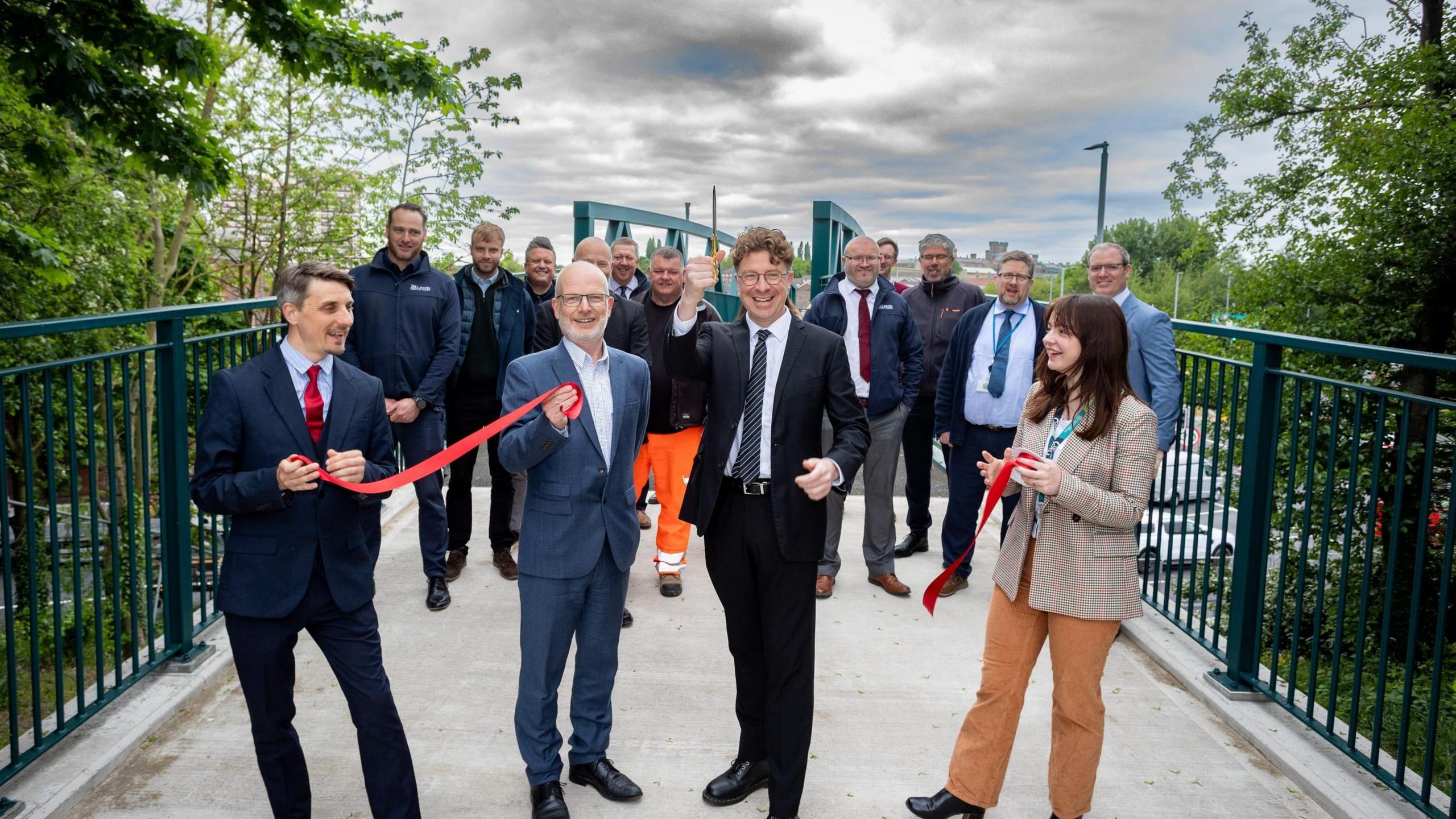 Four smartly dressed people hold red ribbons and smile while stood on a footbridge. A larger group of people, almost all of them men, standing behind them in the background.