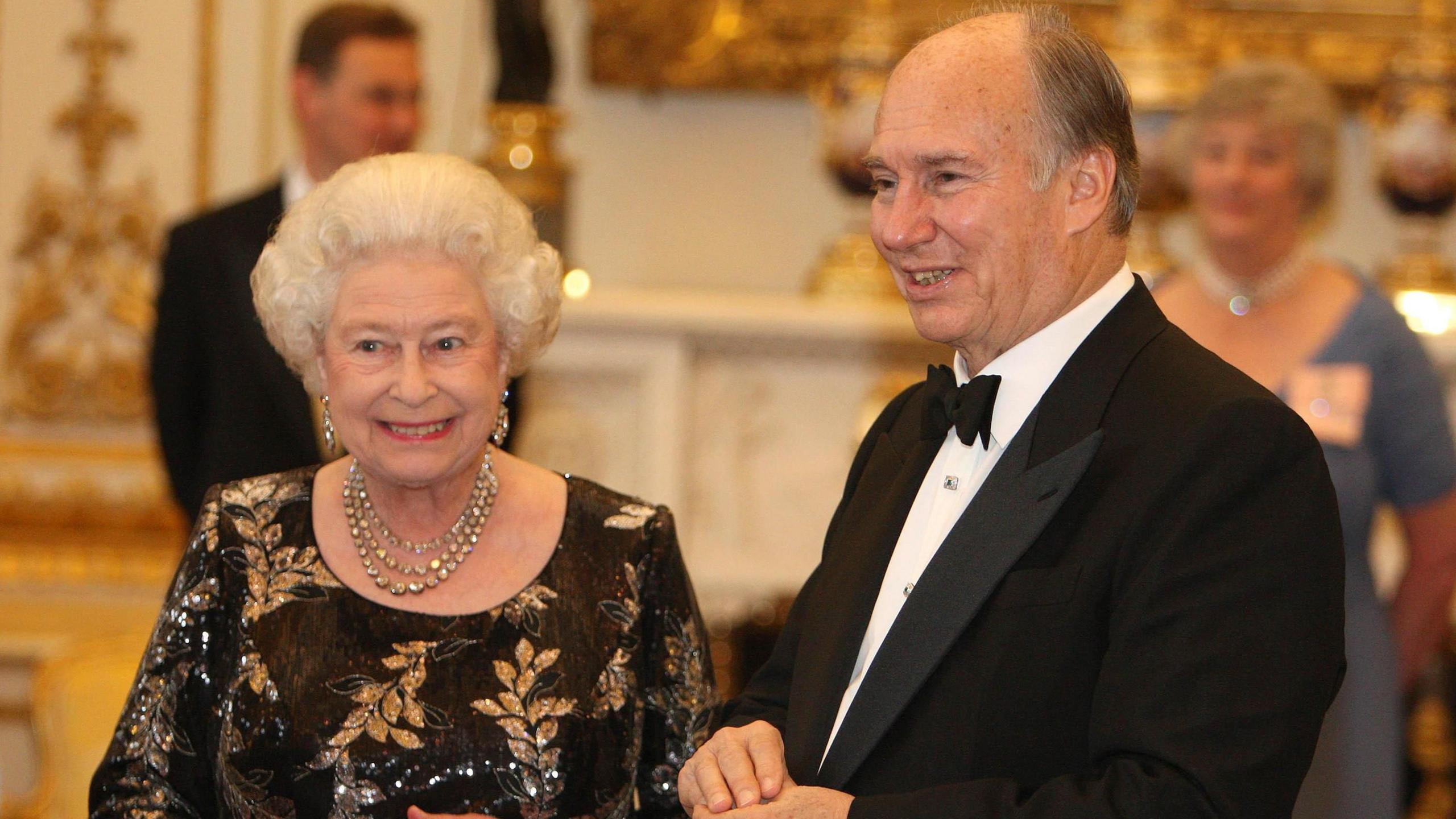 Queen Elizabeth II wearing a black dress with golden leaf patterns standing next to the Aga Khan wearing a black tuxedo