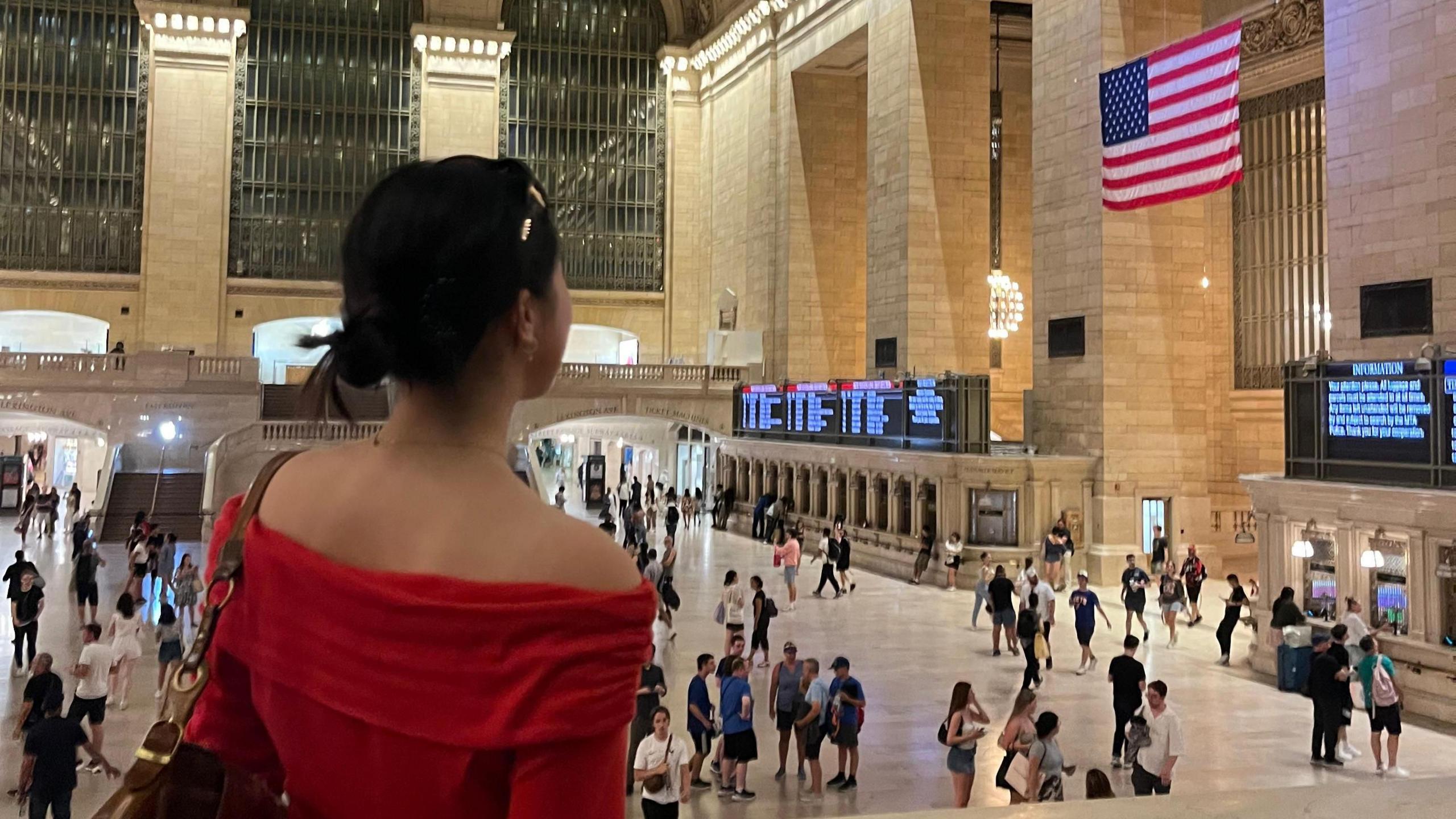 Tian is in a train station. She is looking away from the camera looking at an american flag on the wall. She is wearing a red top.