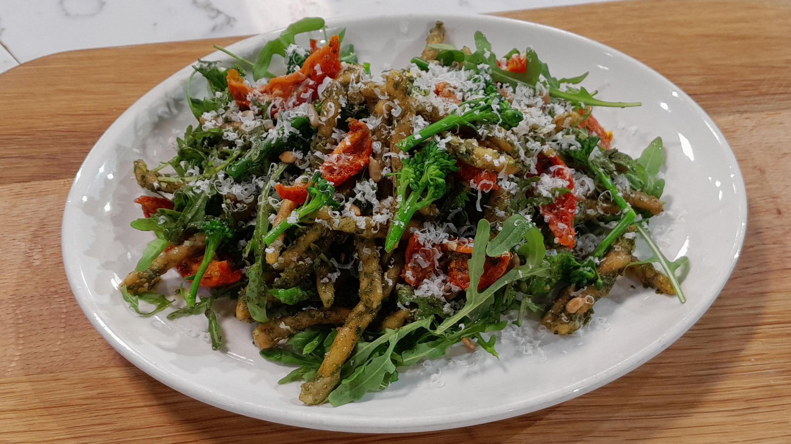 A plate of Anna Haugh's homemade trofie pasta with rocket pesto, garnished with rocket, broccoli and sun blushed tomatoes. The plate is sitting on a wooden board.