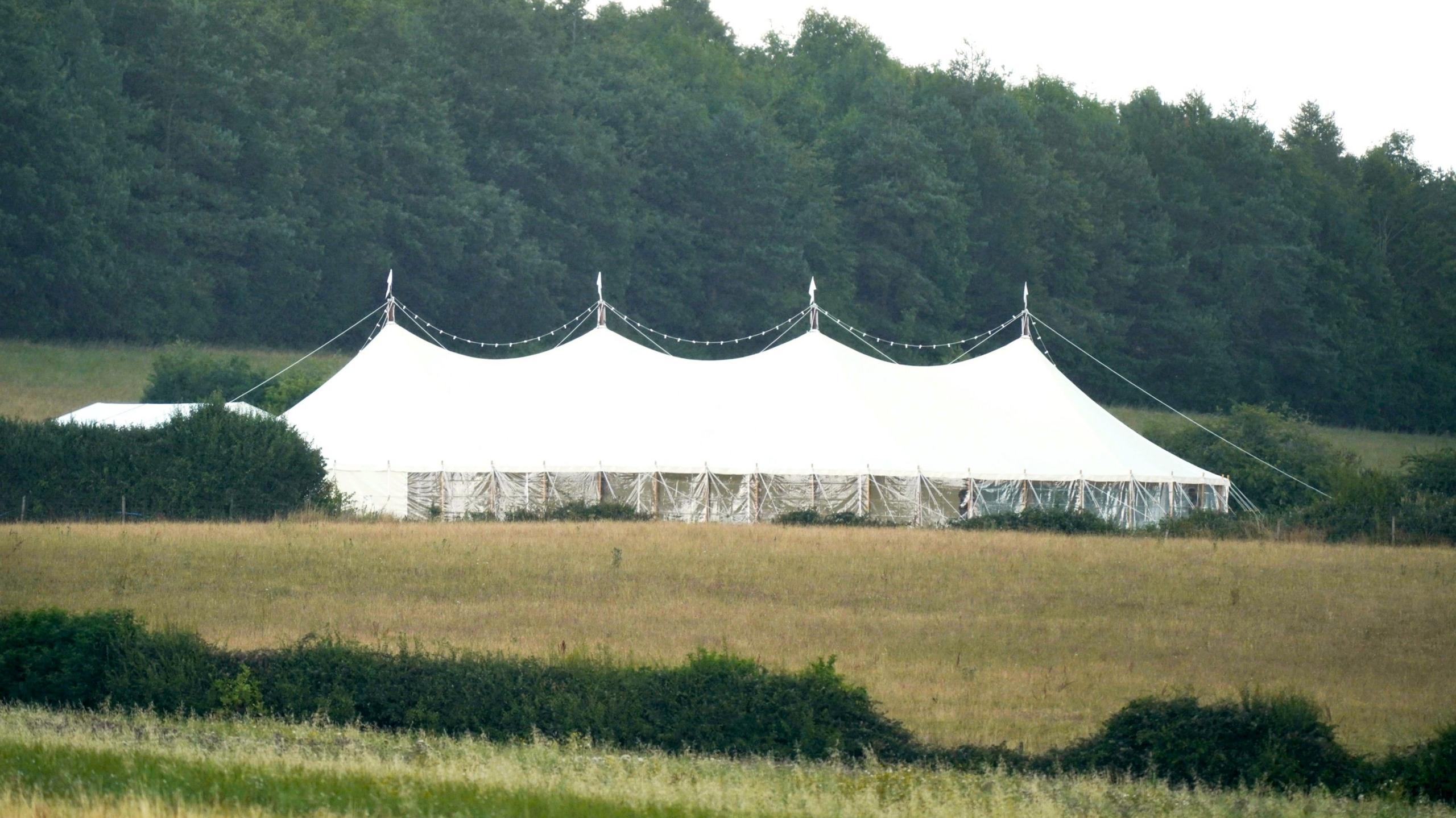 A view across a field showing four peaks of a large white marquee set up in the countryside. There are fairy lights draped in a string over the top and in the background there is a row of large, dense green trees.