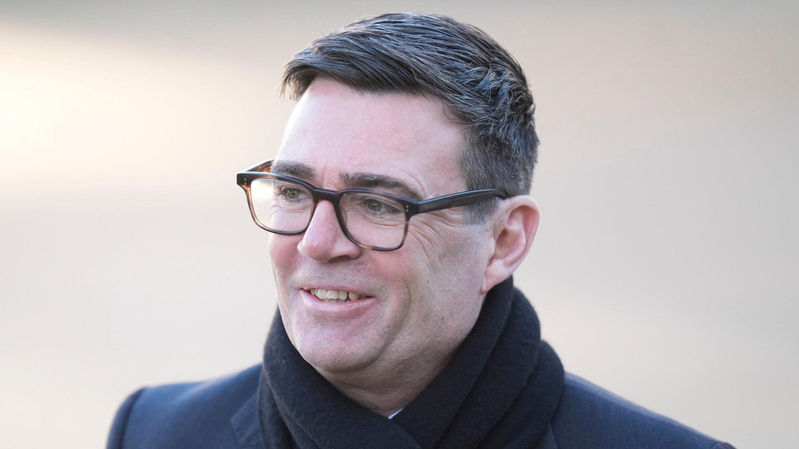 A head shot of Greater Manchester Mayor Andy Burnham. He has short dark hair and is wearing glasses, a dark scarf and a dark coat. He is looking to one side and is smiling.