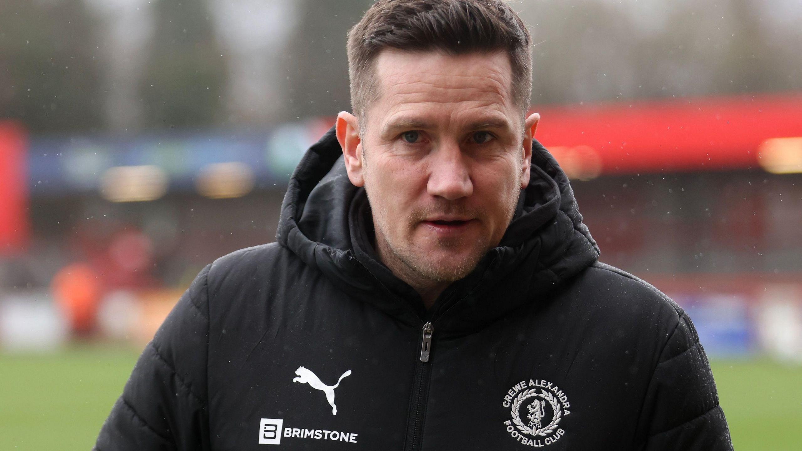 Crewe manager Lee Bell walks towards the dressing room after a game in his black club puffer coat