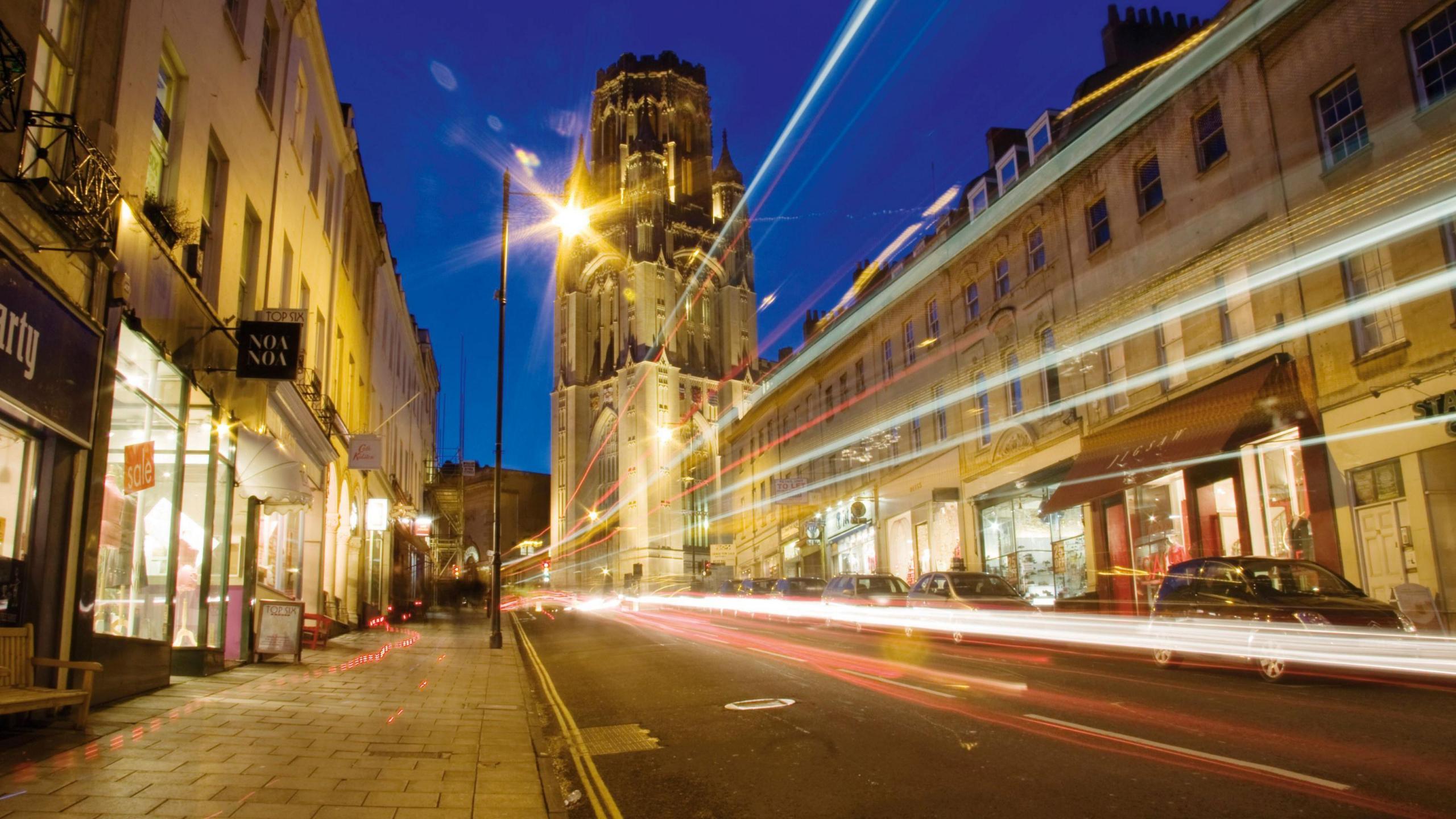 An image looking up Park Street in Bristol at night. The picture is taken using long exposure settings so headlights and brake lights from vehicles appear as long lines of red or white light. Either side of the road the illuminated fronts of shops are visible, and the Wills Memorial Building can be seen at the top of the street