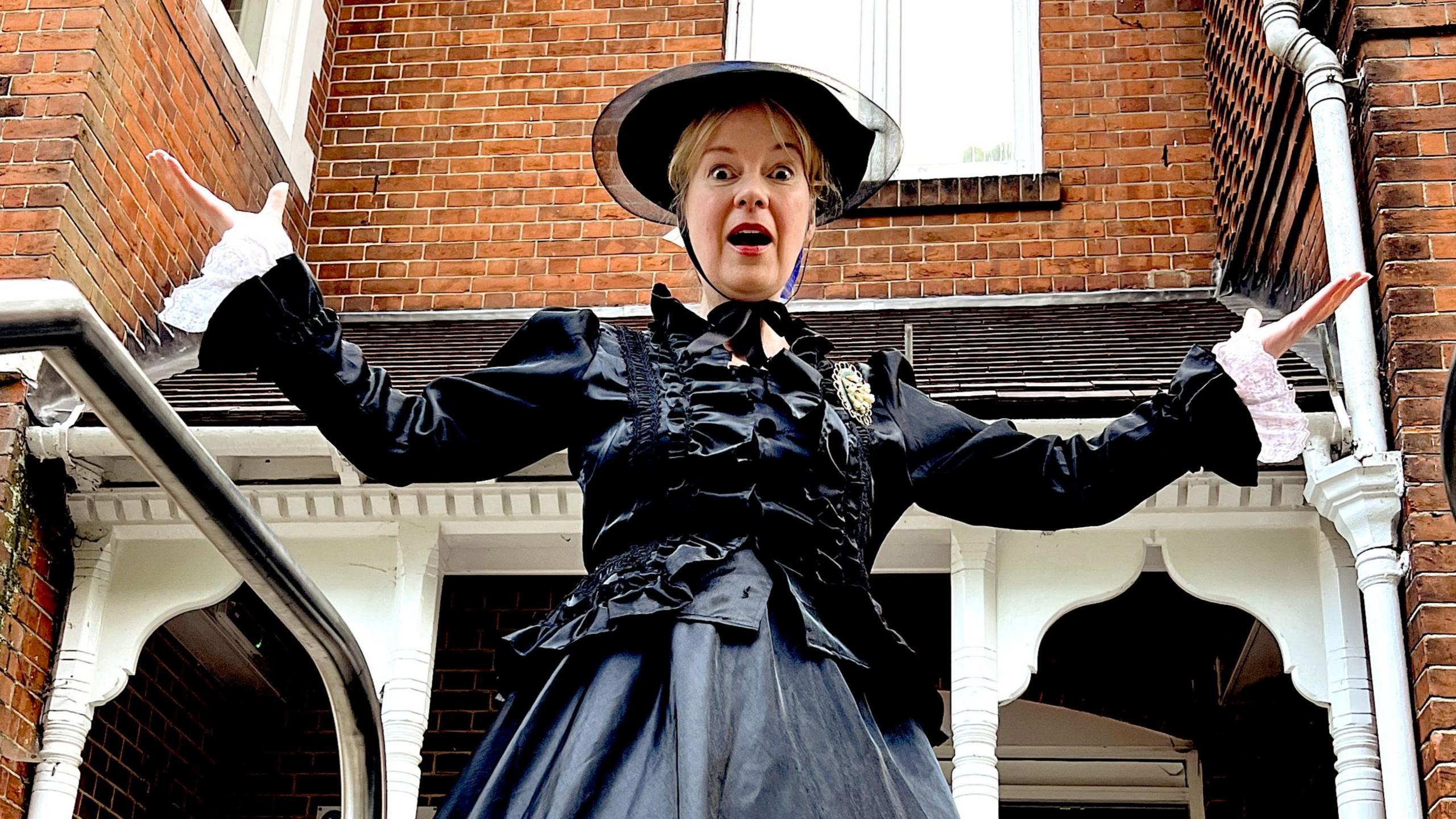 A woman dressed as Mary Shelley in a long black dress is looking shocked with her arms outstretched. She is standing outside a brick house with a decorative white wooden porch. She is looking down at the camera.