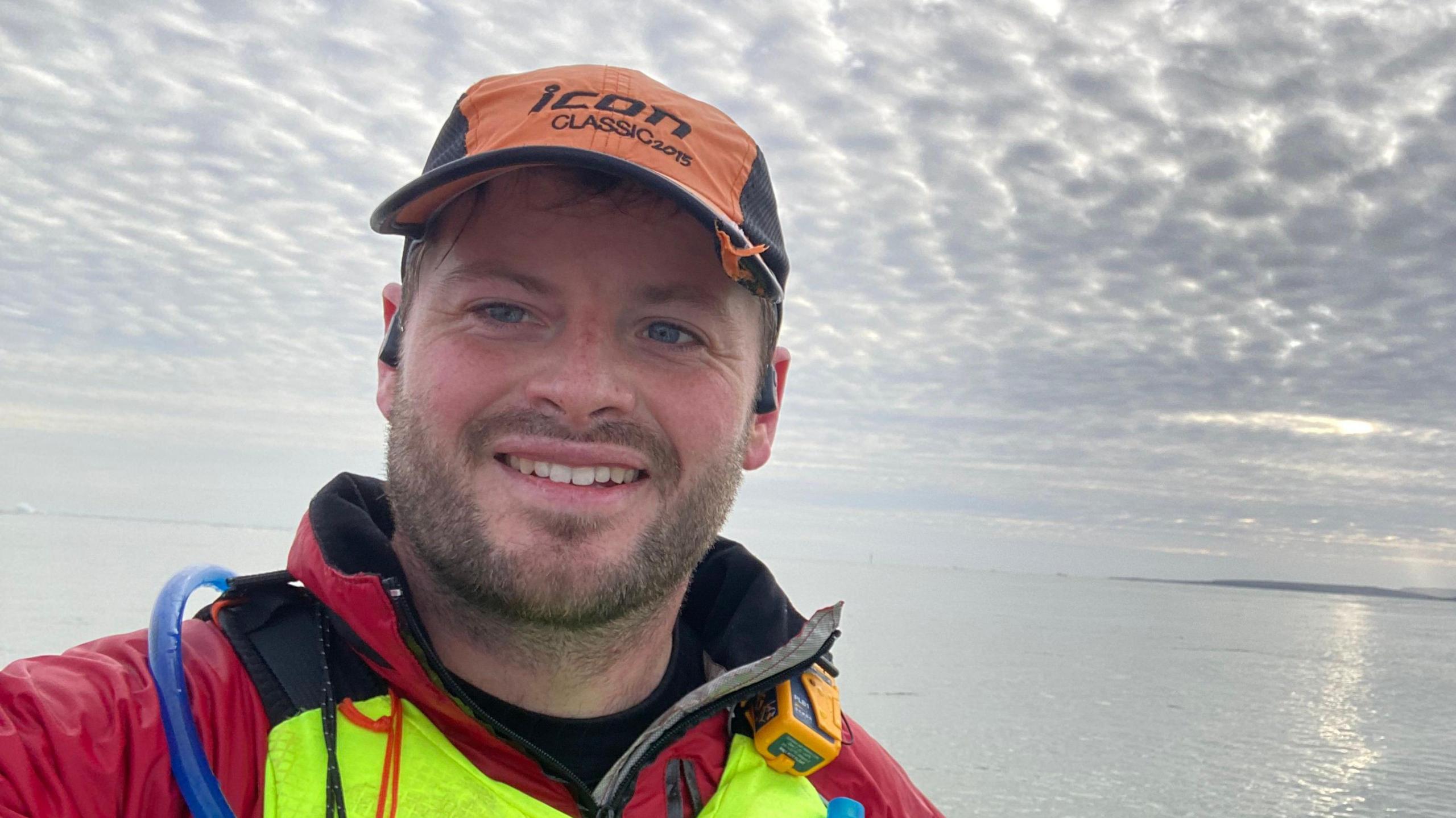 A man in a red jacket and yellow lifejacket smiling at a camera whilst on a kayak at sea