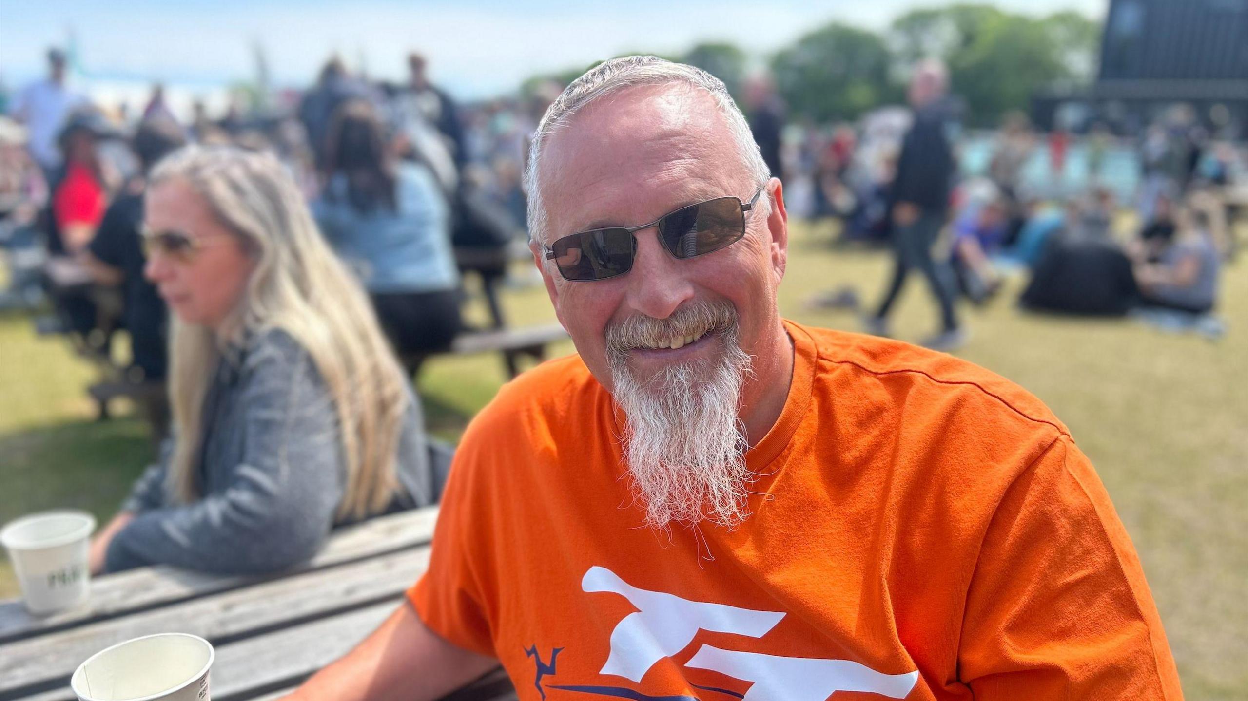 Derek Adams wears a bright orange TT t-shirt, with white writing. He wears smell dark sunglasses and has a long white beard. He sits at a bench holding a paper cup. You can see other spectators in the background.