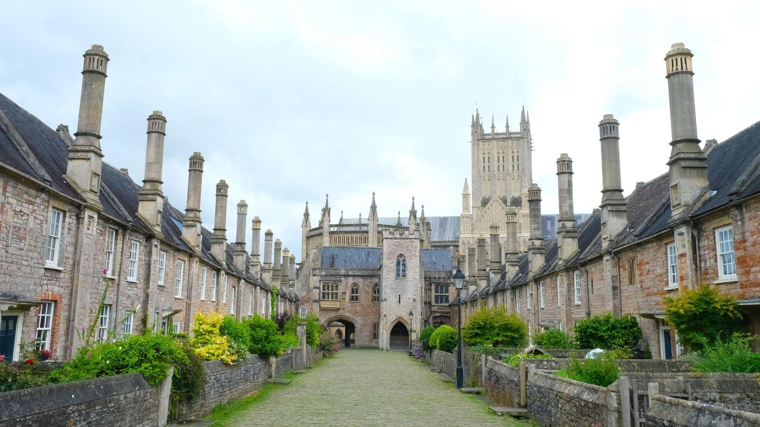 Vicars' Close with Wells Cathedral in the background, the close is paved with grey cobbles with grass growing through them. On either side are rows of terraced houses with large chimneys extending from the roofs. 