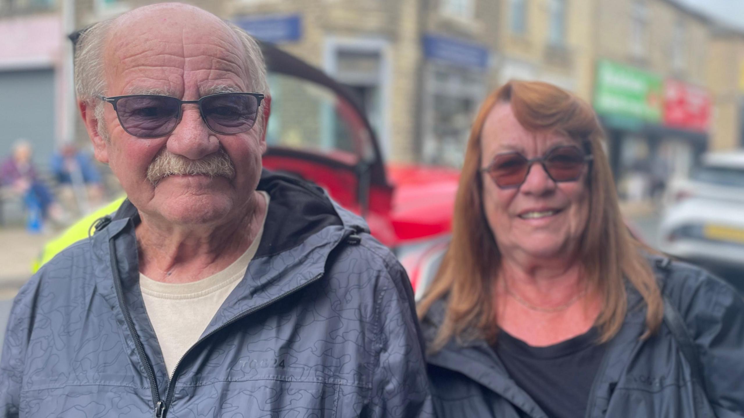 A man wearing dark tinted glasses with a moustache is stood to the left of the picture.
A lady wearing tinted glasses and a grey coat is to the right of the picture.
Behind them is a road with cars on it.