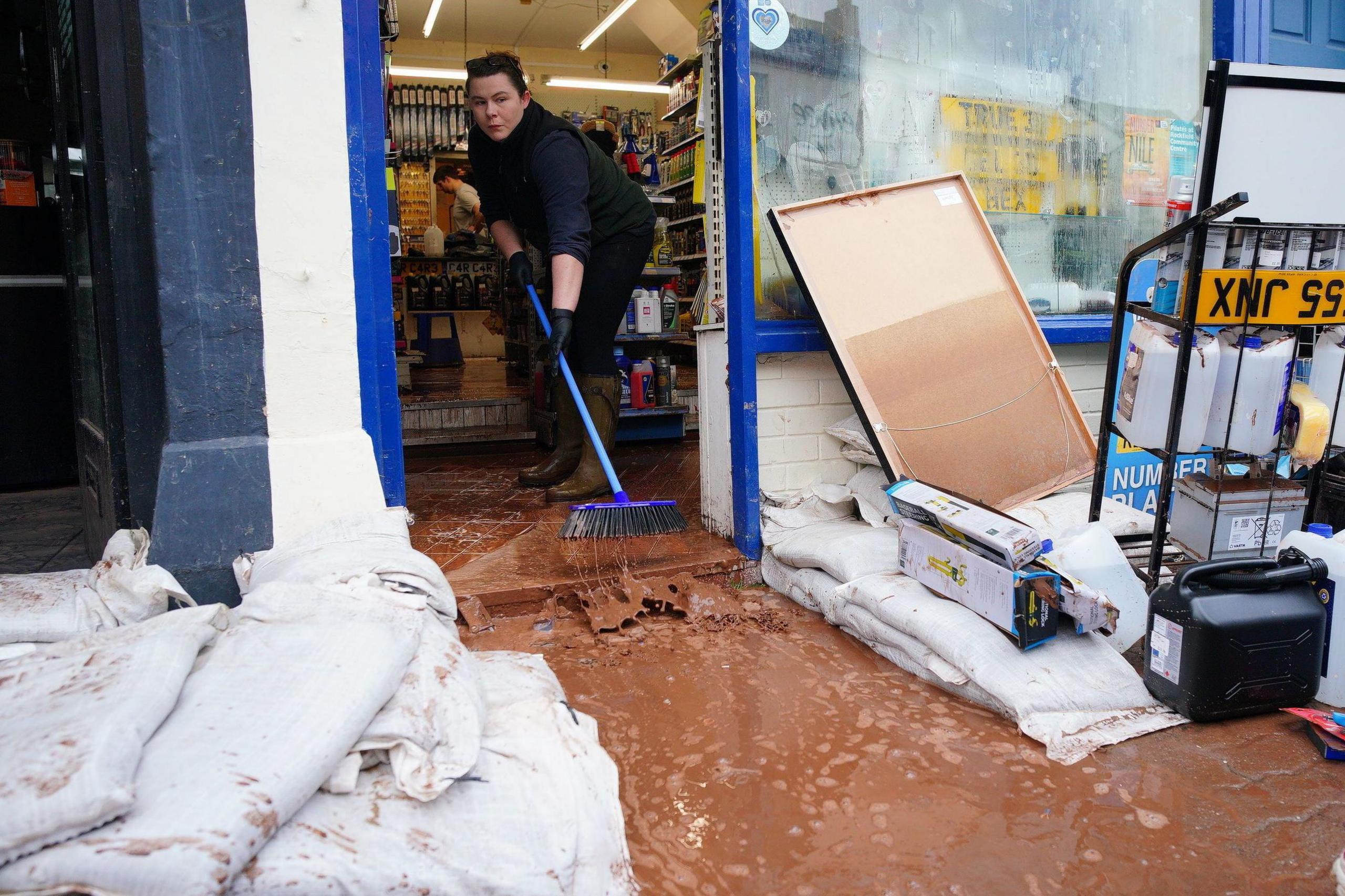 Woman sweeps brown muddy water out of a shop door with a gap made in sandbags over the front step