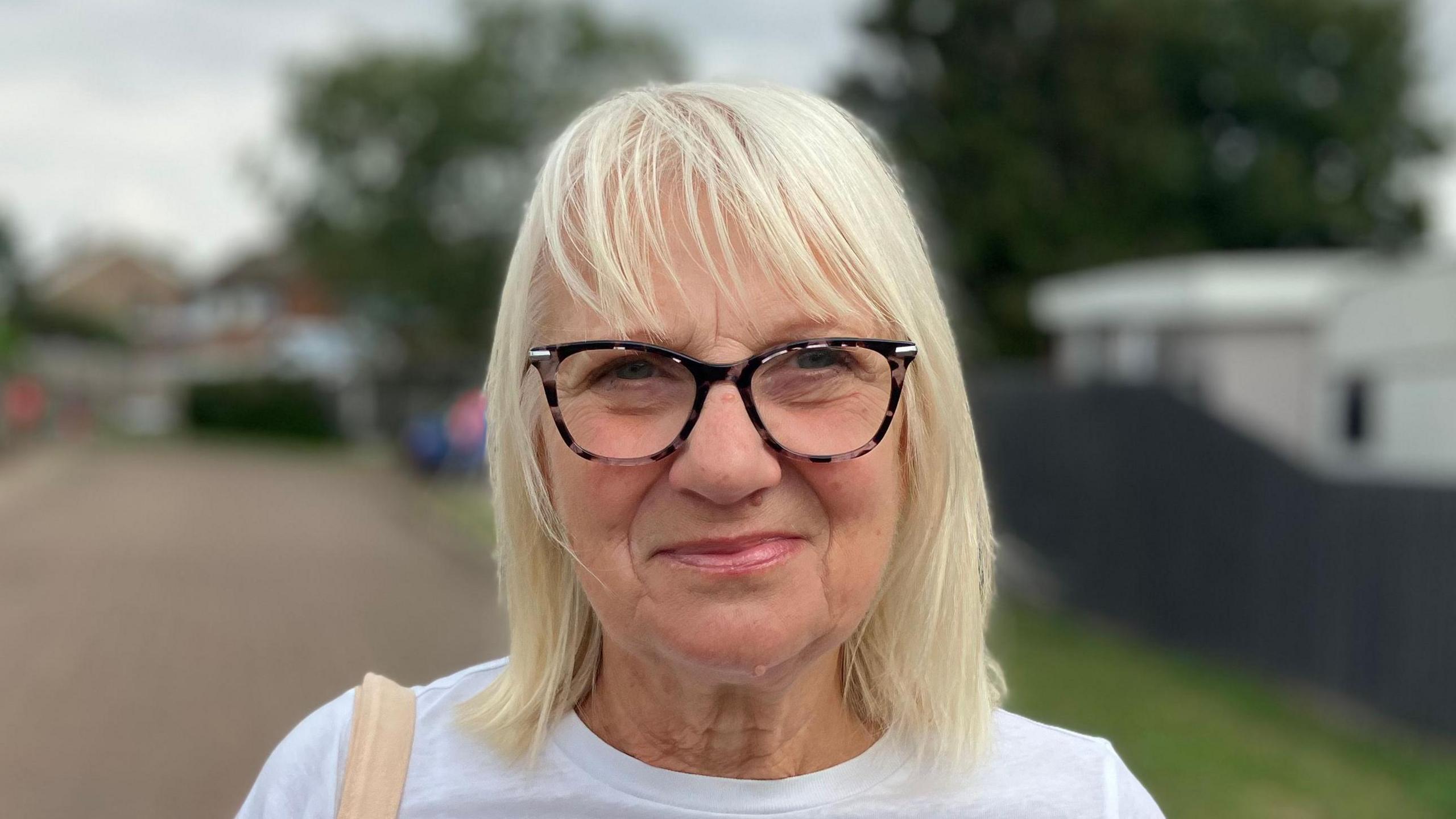 Jane Carrington smiles at the camera. She has shoulder length white blonde hair and wears glasses and a white T-shirt. A bag strap rests over one of her shoulders.