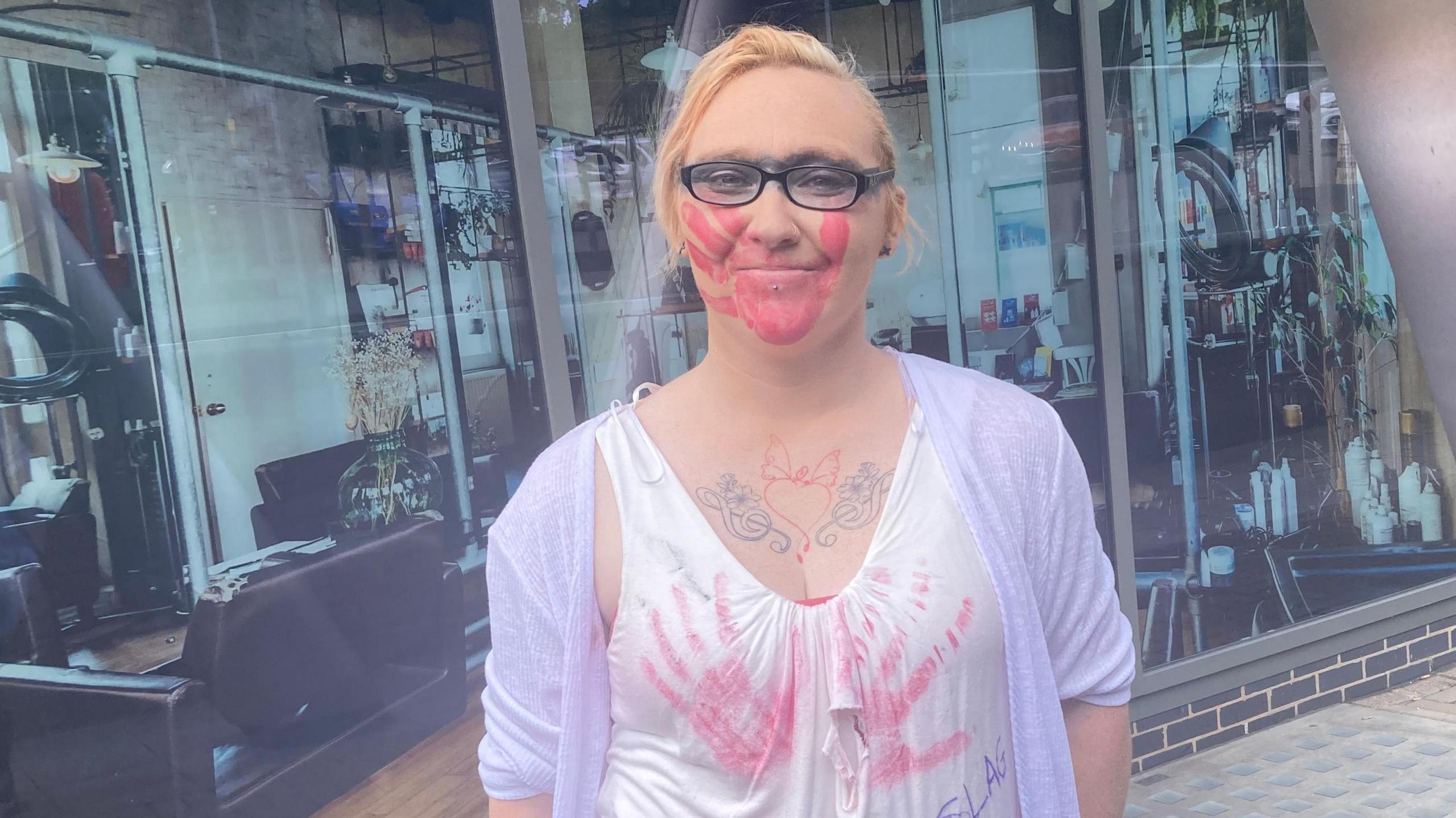 A blonde woman wearing glasses with a line of black face paint covering her eyes and a red hand mark over her mouth. She also has two red handprints on her top over her breasts.