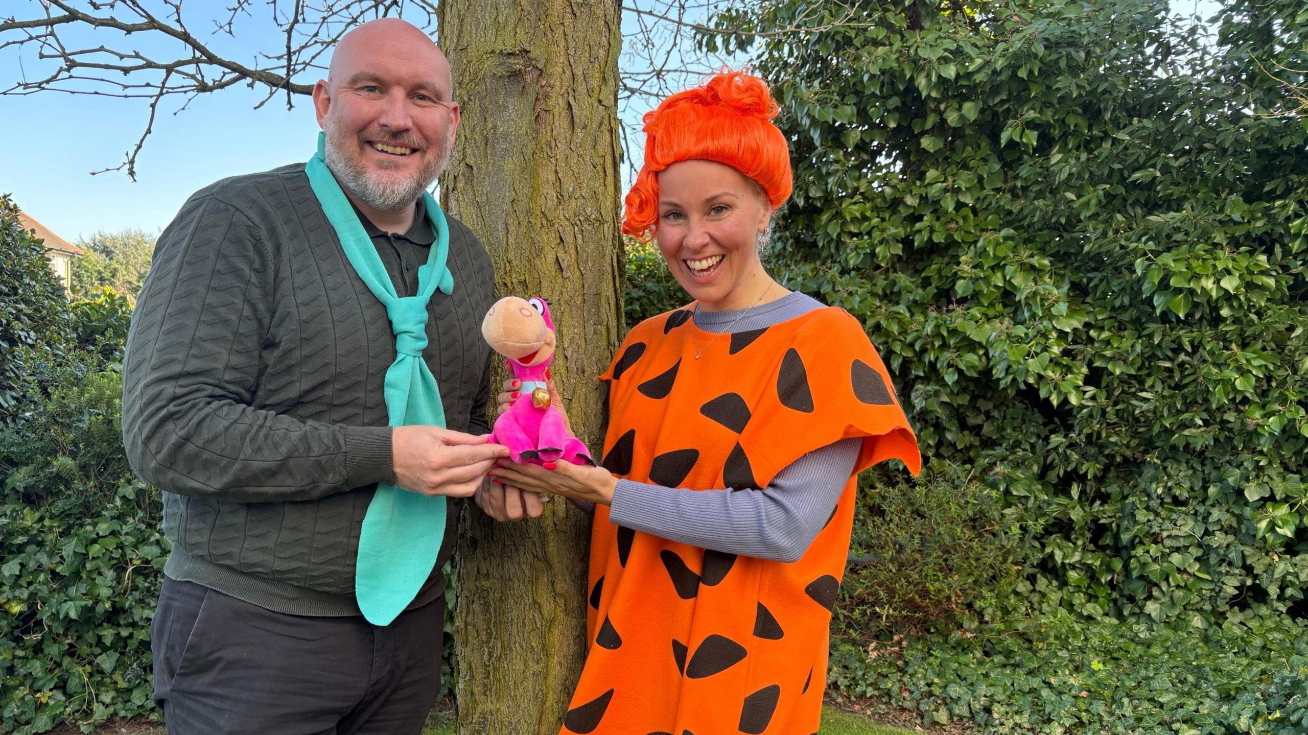 A man and woman dressed as the popular cartoon characters from The Flintstones. They are both smiling at the camera and standing by a tree 