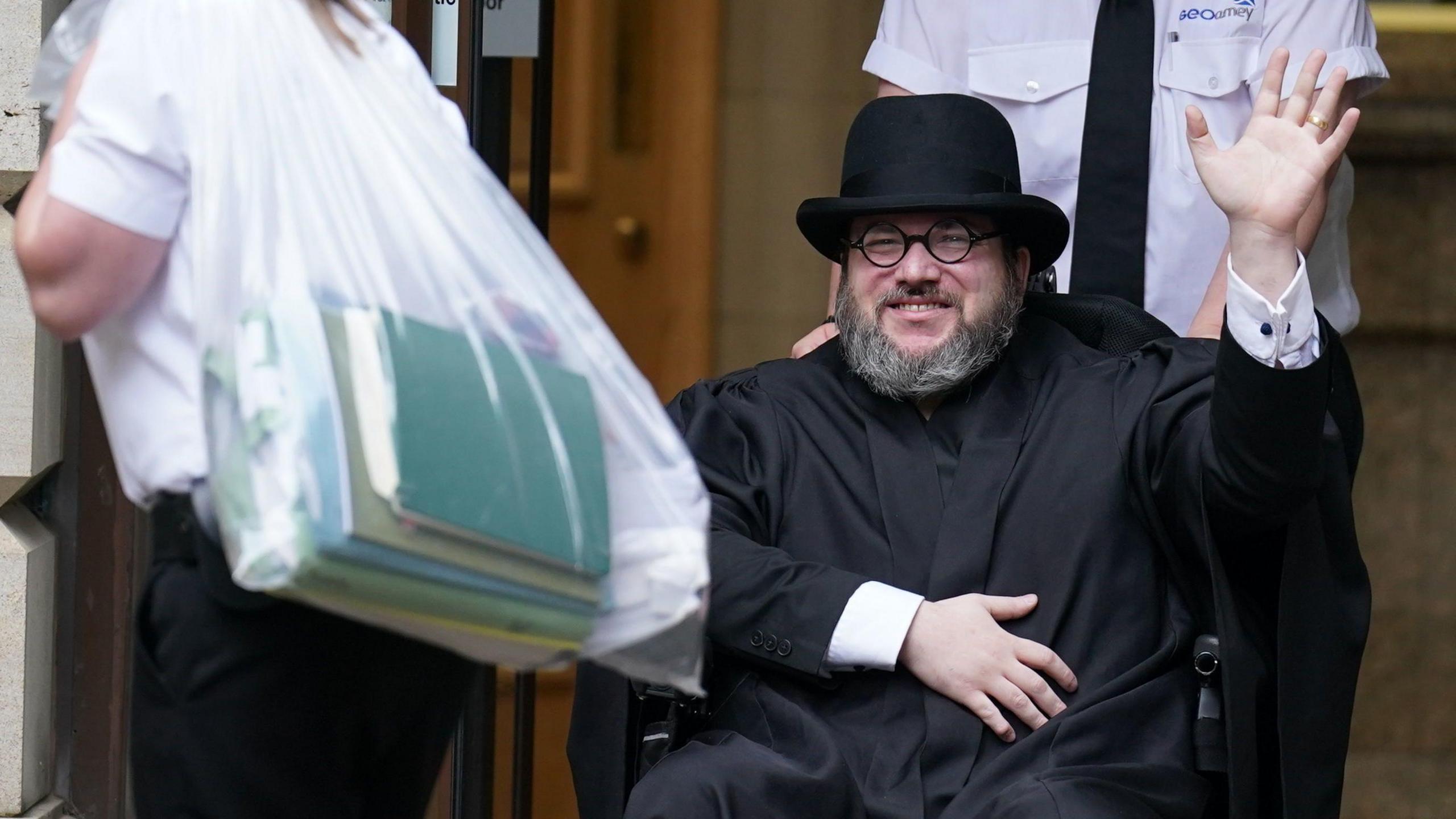 Nicholas Rossi, sitting in a wheelchair wearing a black silk gown and black Homberg hat, waving to the camera while being wheeled out of court by a guard