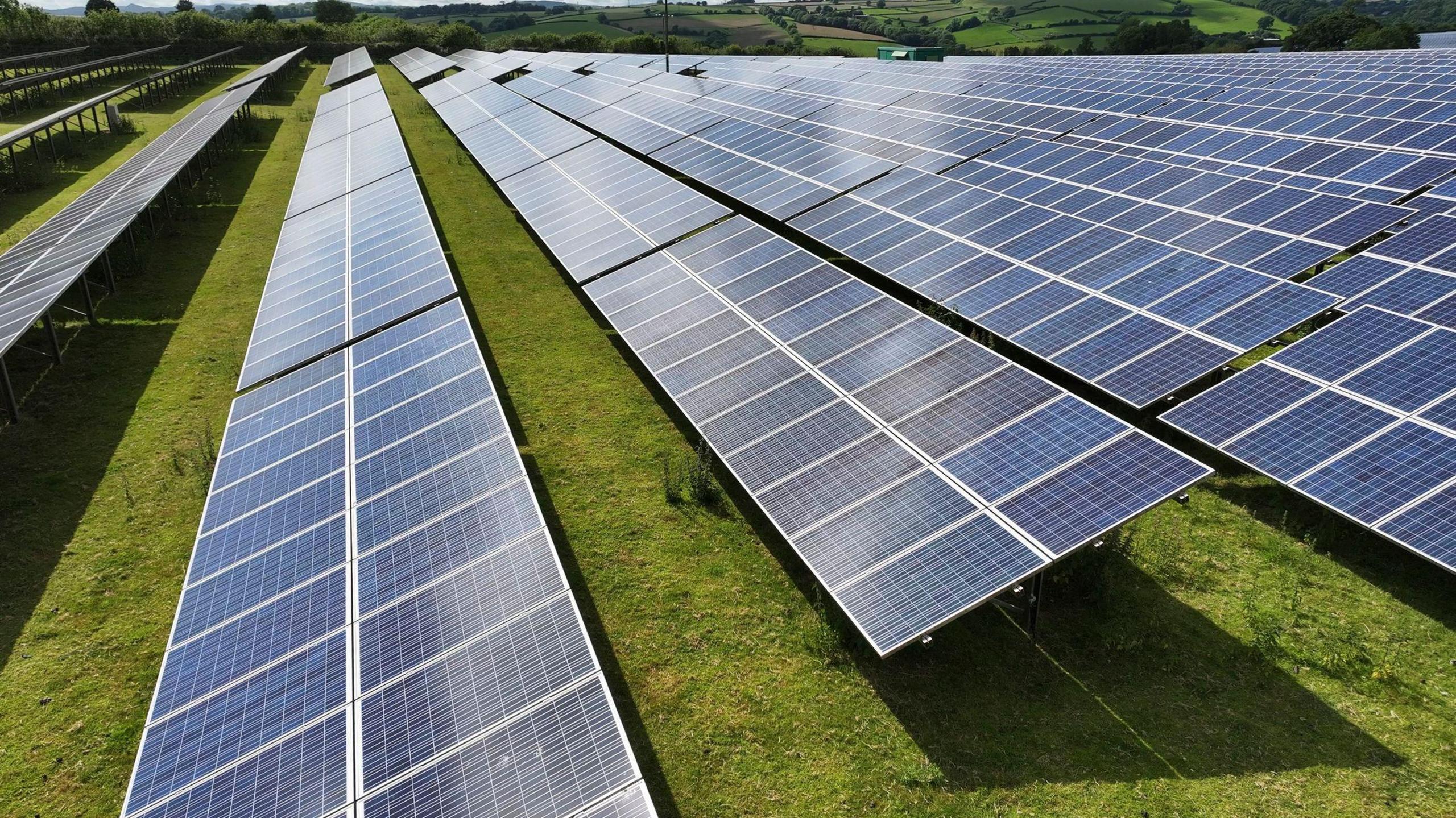 Rows of solar panels fill a grassy field, with a view of the countryside in the distance.