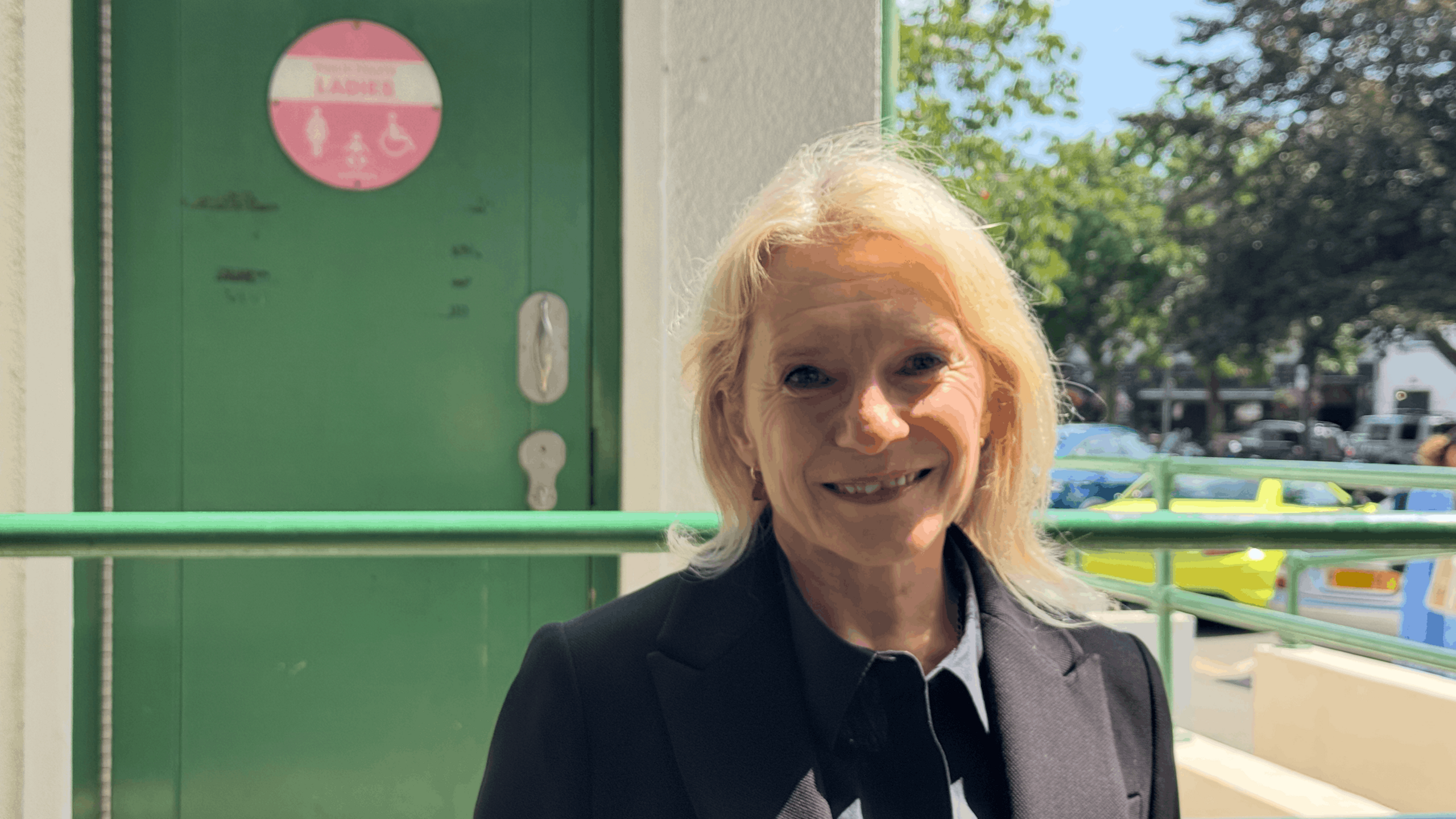 Senator Elaine Millar wearing a dark jacket, standing outside a public toilet entrance, with a green accessible restroom door visible in the background.