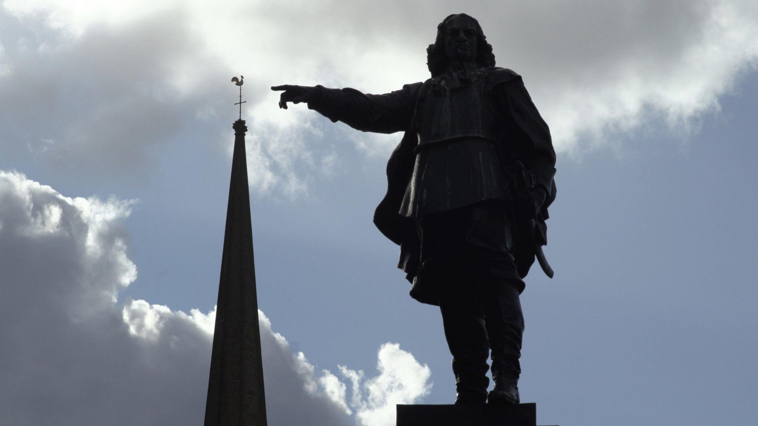 A statue, pictured in sillhouette and from below, showing a man in a long jacket pointing at the spire of an adjacent building. Blue skies and white fluffy clouds are visible in the background. 