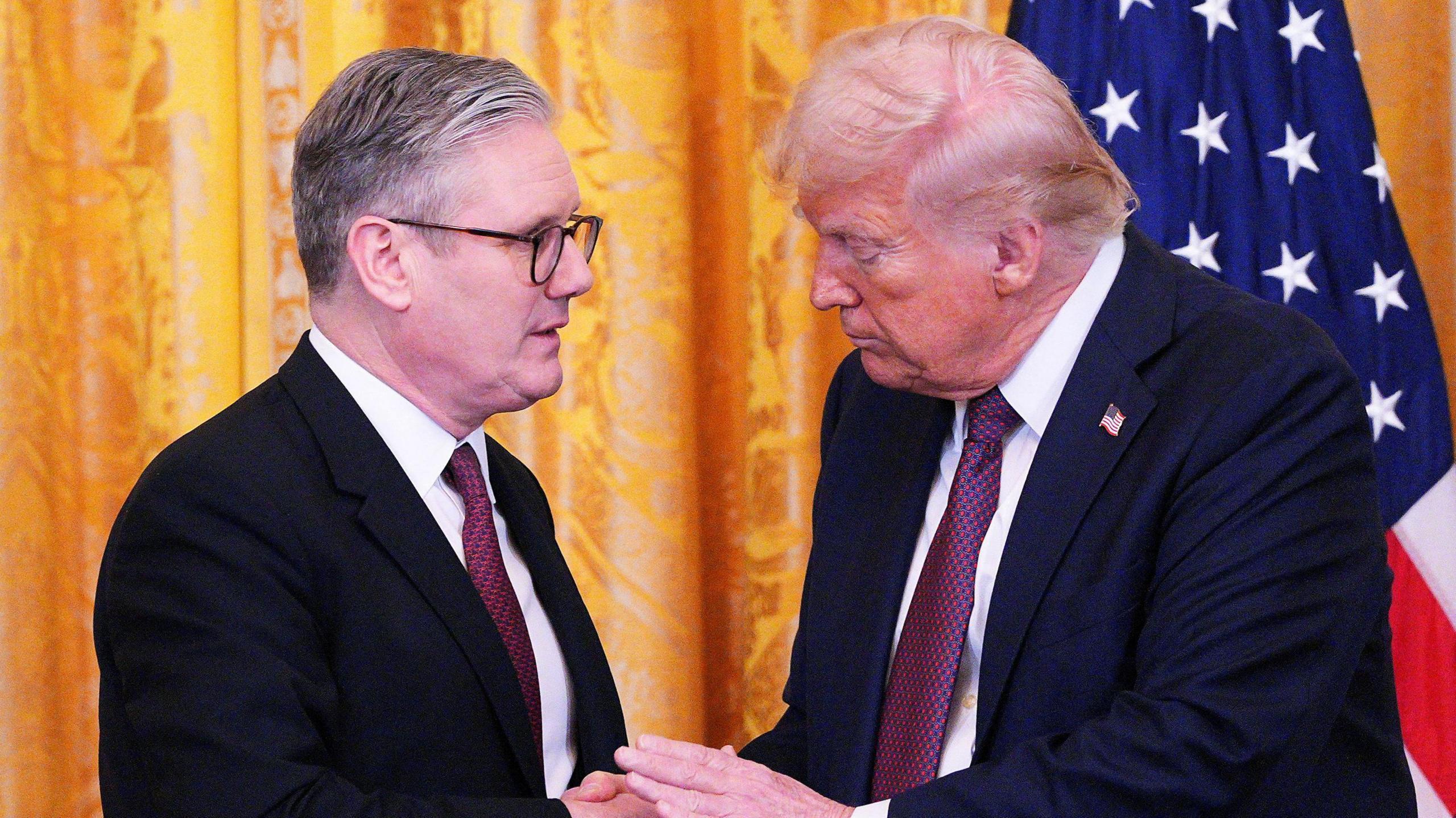 UK Prime Minister Keir Starmer on the left in a black suit and red tie shaking hands with Donald Trump on the right in a navy suit and red tie with an American flag pin on his lapel. They're standing in front of a yellow curtain with the US flag behind Donald trump on the right hand side