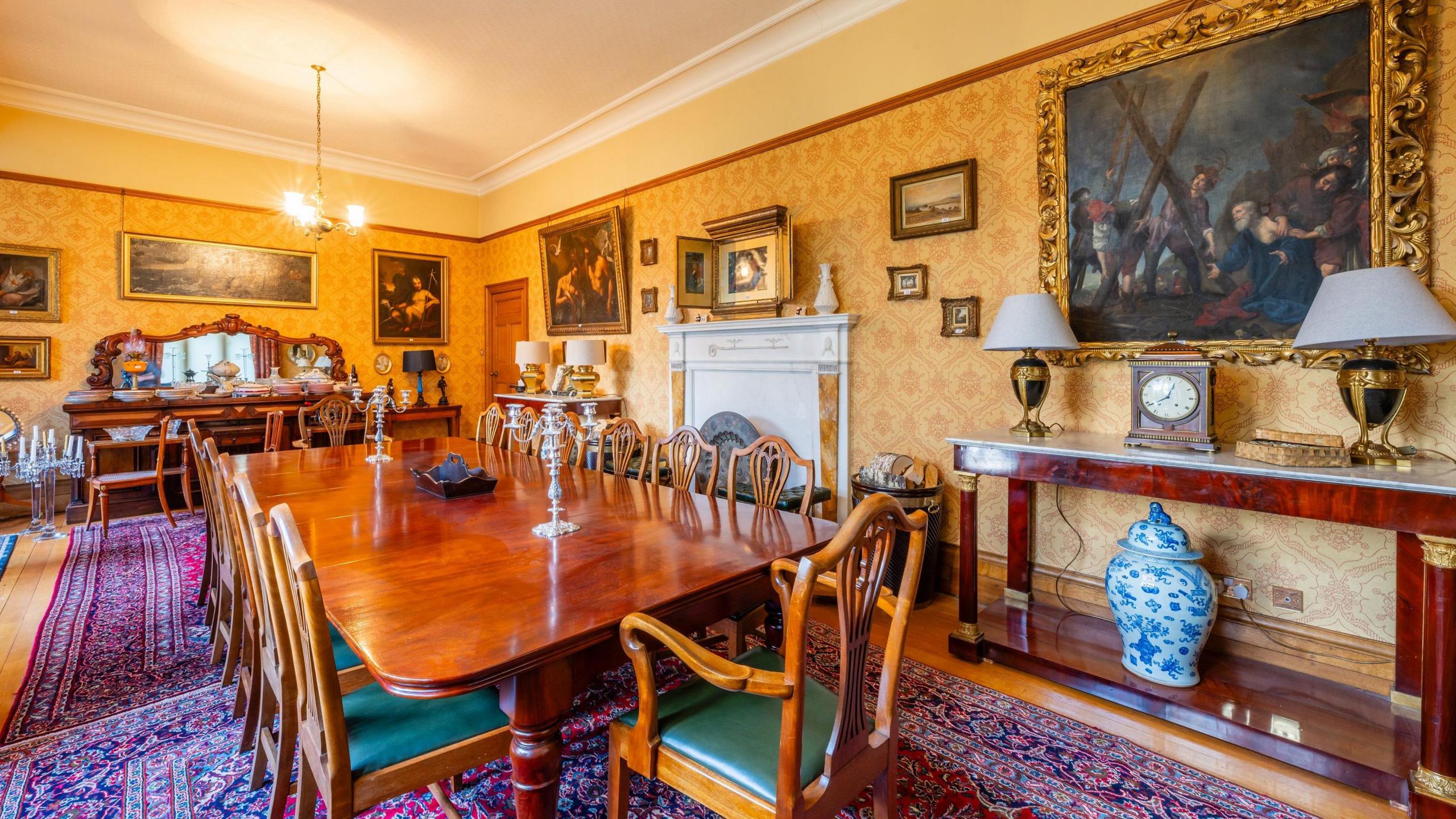 The dining room in the Bishop's House in Newry.  It is an ornate wallpapered room with several framed paintings, a marble fireplace, a long wooden dining table and other furniture and ornaments. 