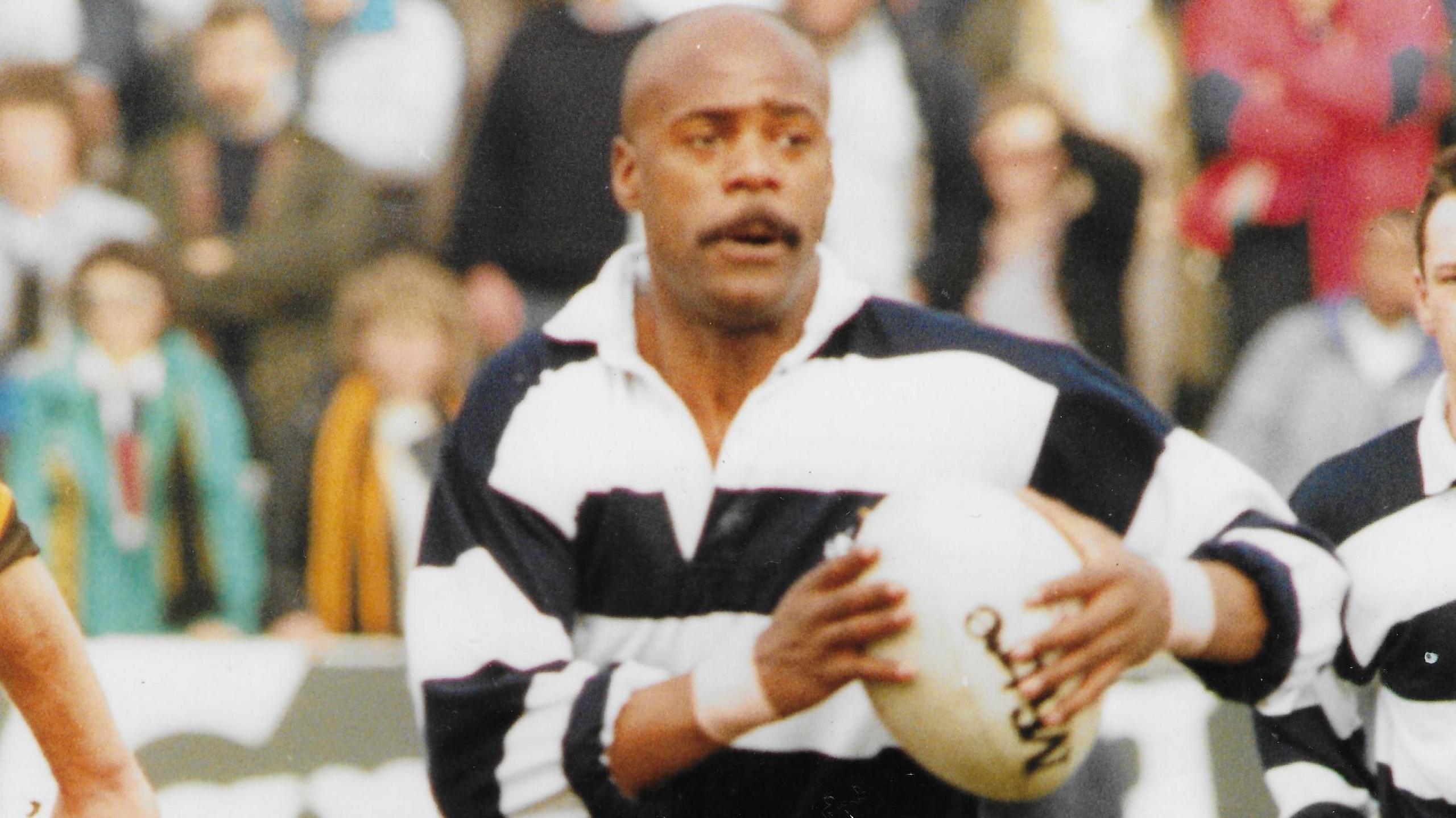 Ralph Knibbs playing rugby. He has a moustache and is wearing a black and white striped long sleeve collared shirt. He is holding a rugby ball and is looking away from the camera, in the middle of play. A crowd of spectators can be seen in the background. 