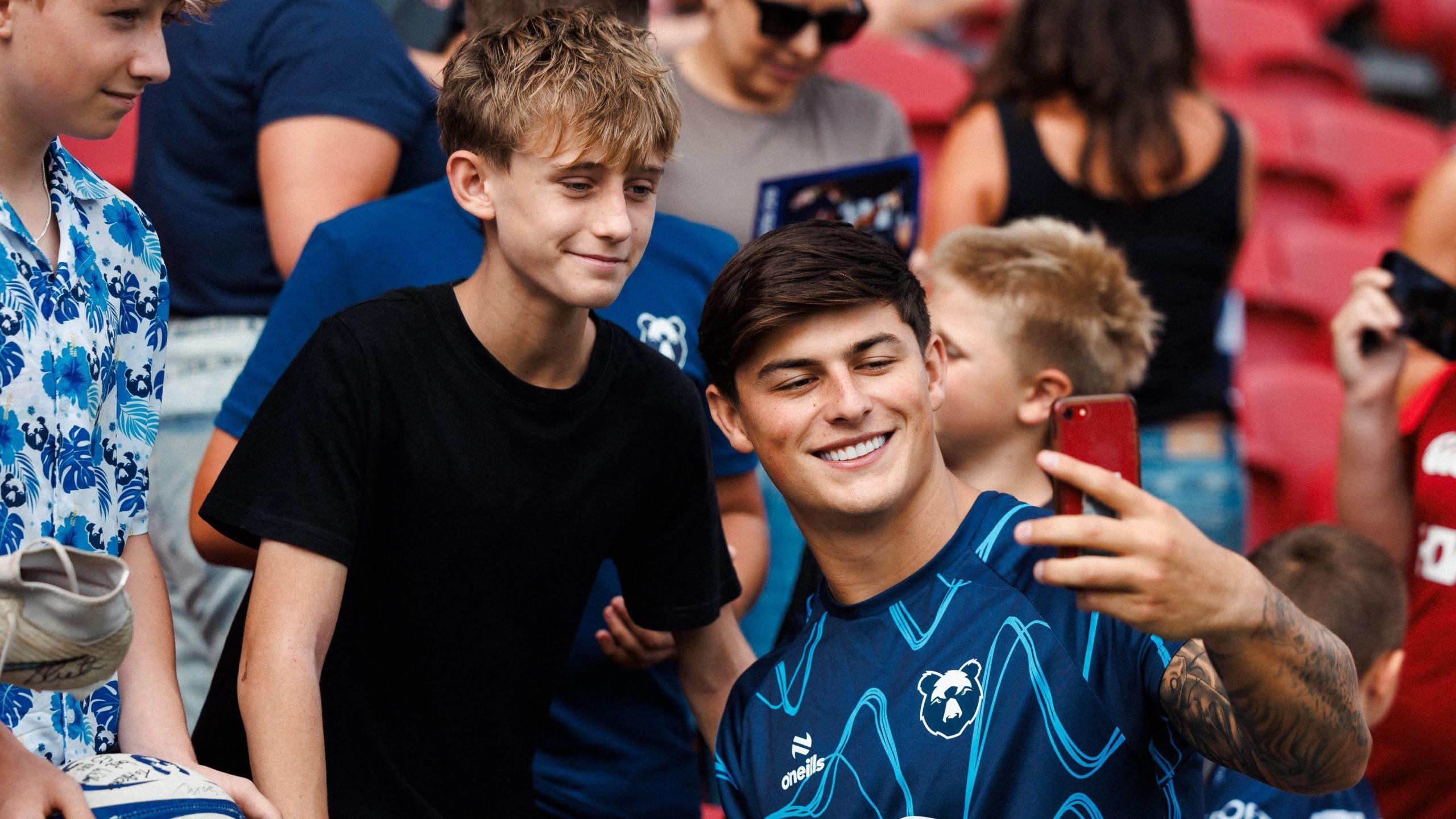 Louis Rees-Zammit, wearing a blue Bristol Bears kit, smiles as he takes a selfie with a young fan during a training session at Ashton Gate. Rees-Zammit is smiling and the phone he is holding to take the photograph is red. The young fan is wearing a black T-shirt and has wavy brown/blond hair.