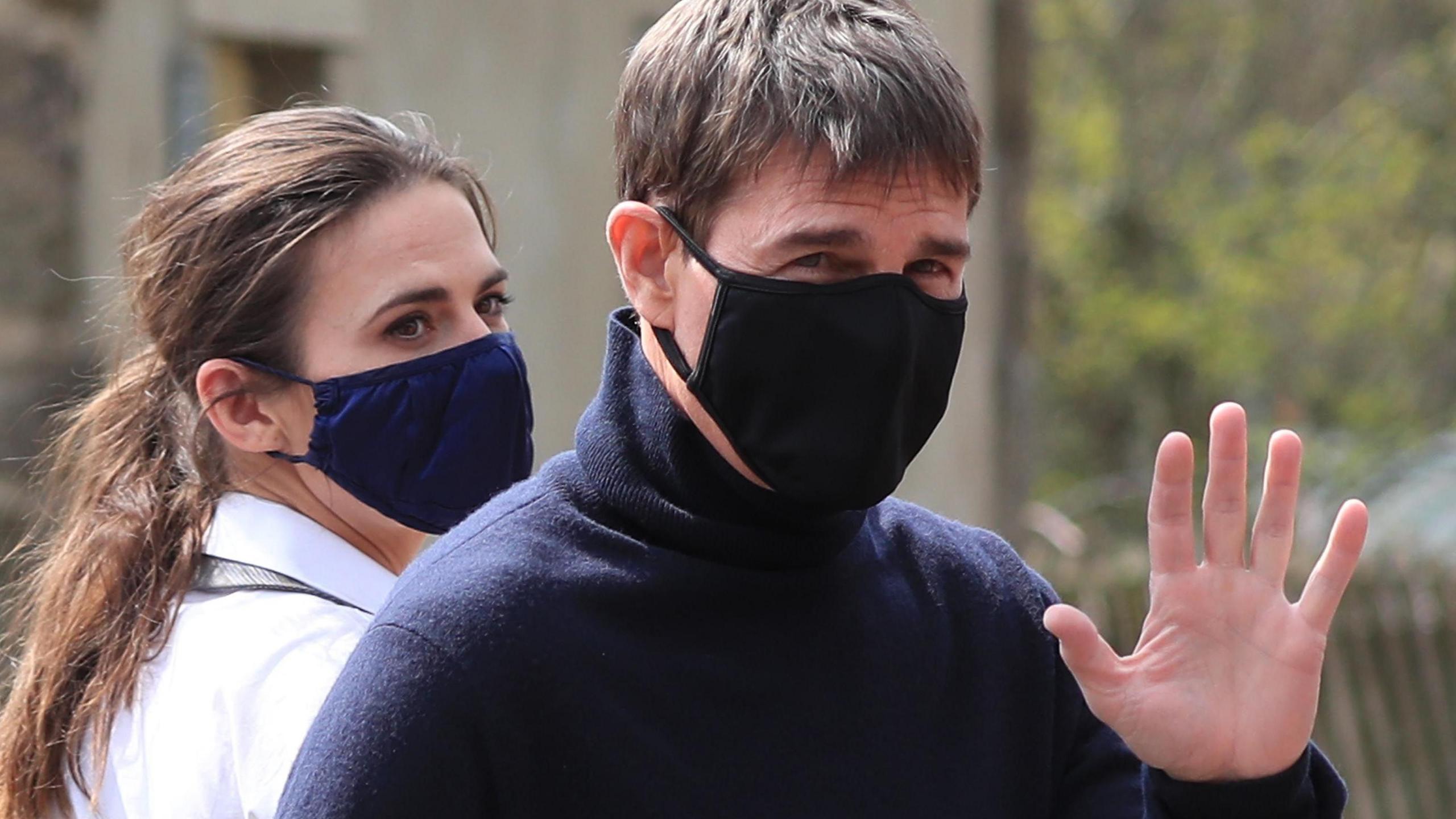 Tom Cruise wearing a dark polo neck and a black Covid-style face mask waves at the camera